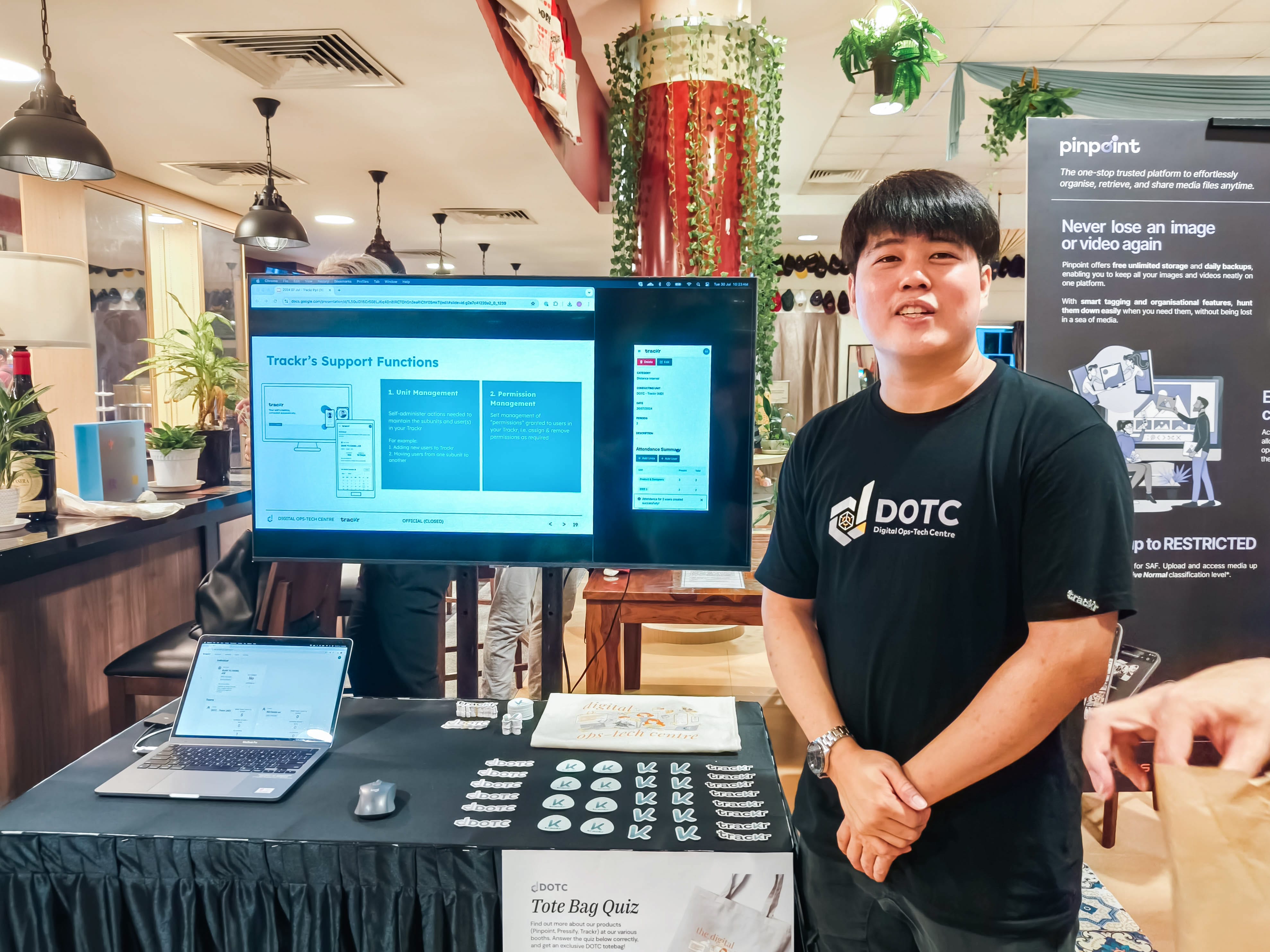 A young man in a DOTC black t-shirt stands at an exhibition booth with a monitor showing Trackr's Support Functions.