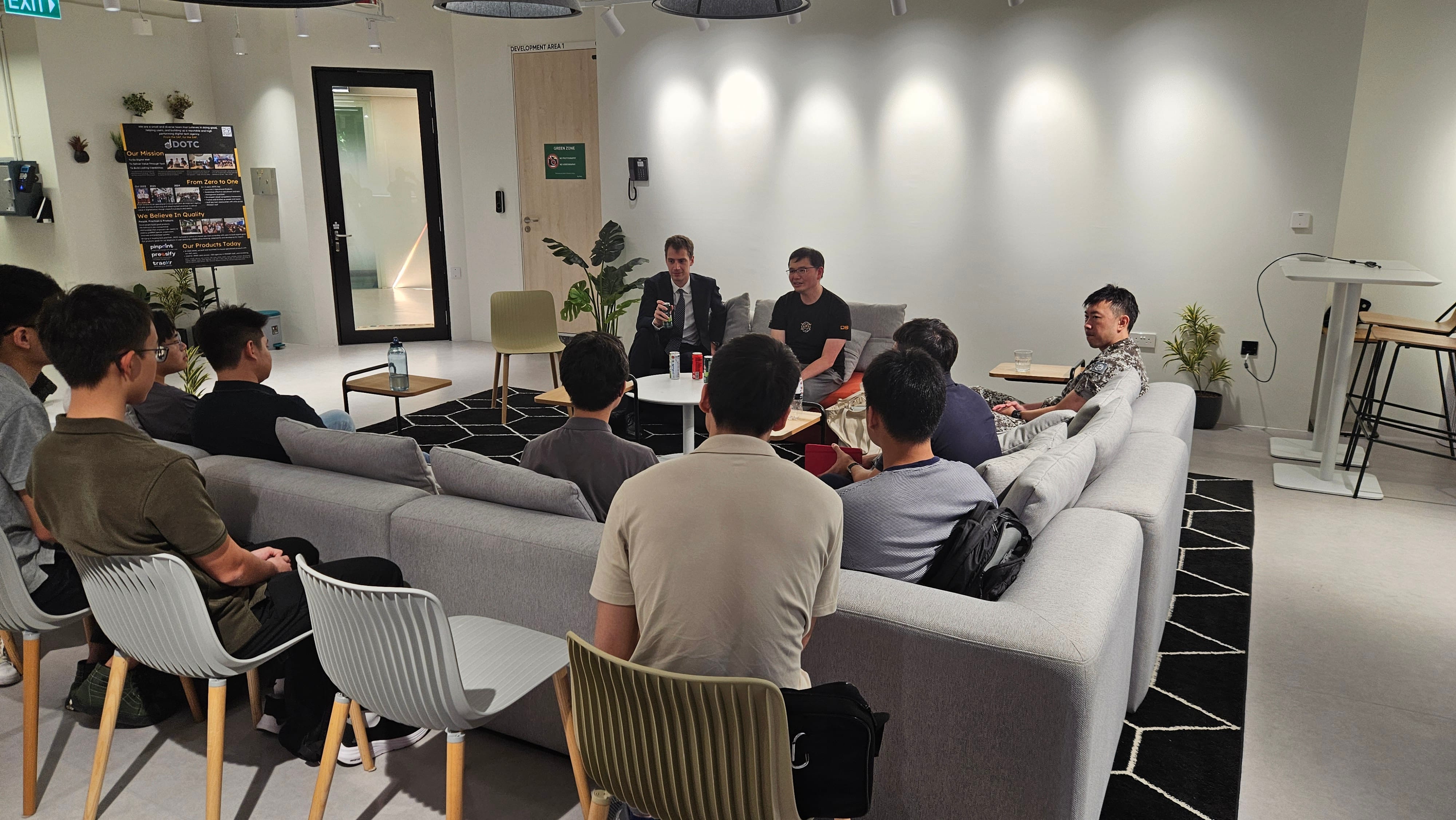 A group of people seated on sofas and chairs in a modern office lounge, listening to two men speaking at the front.