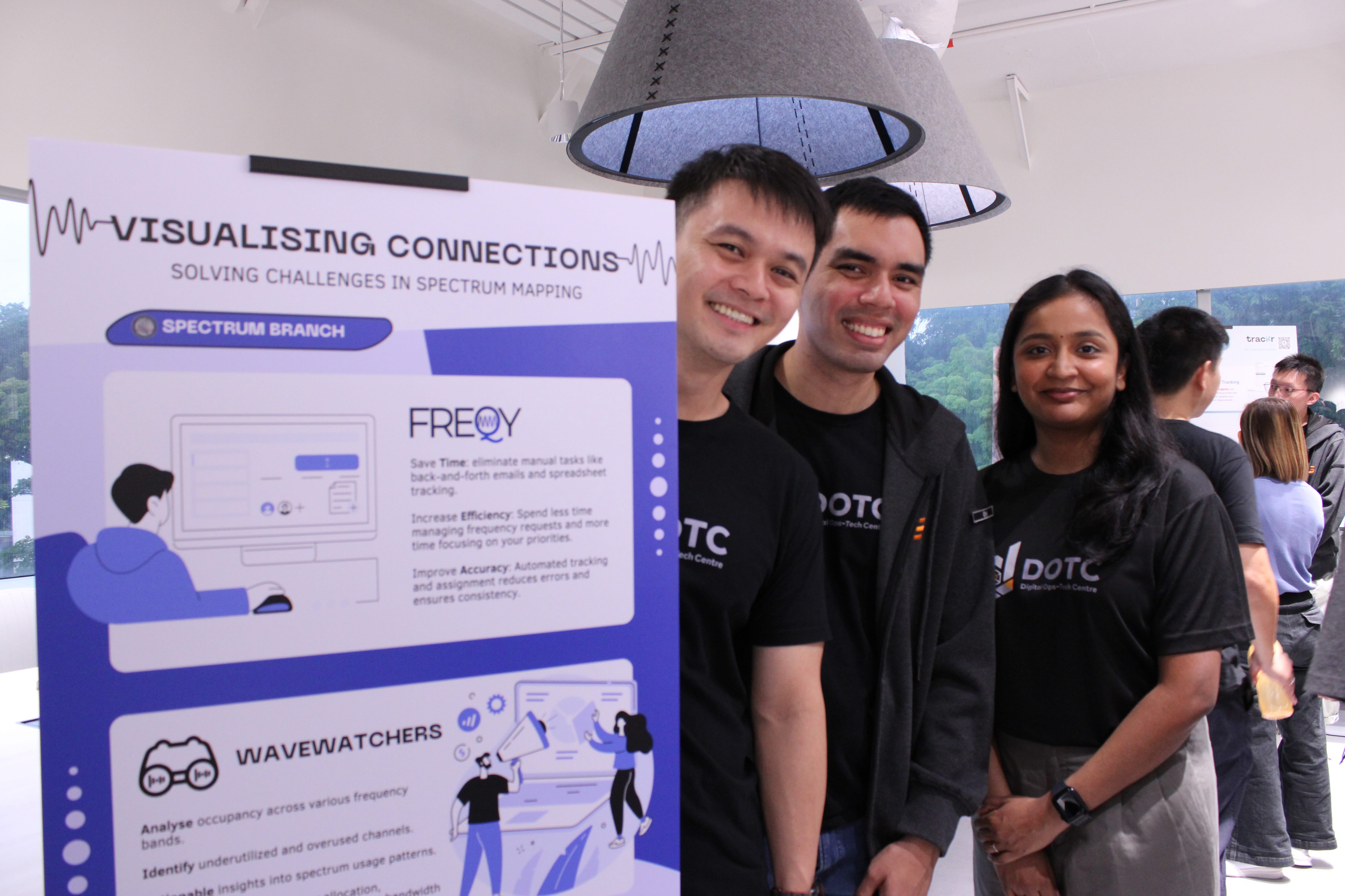 Three smiling people in DOTC shirts stand beside a poster titled Visualising Connections about spectrum mapping.