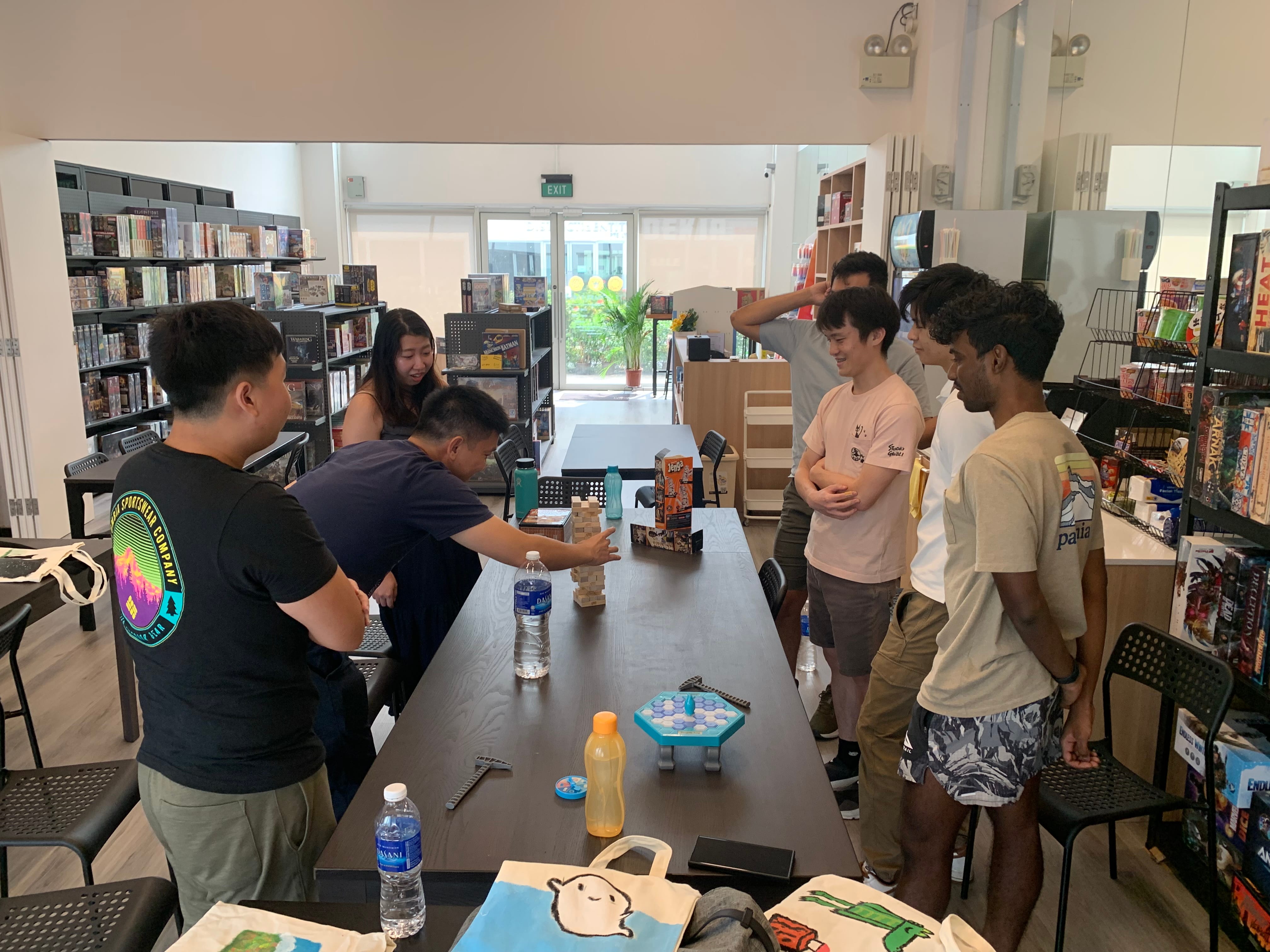 Six people stand around a table playing Jenga in a board game cafe with shelves of games in the background.