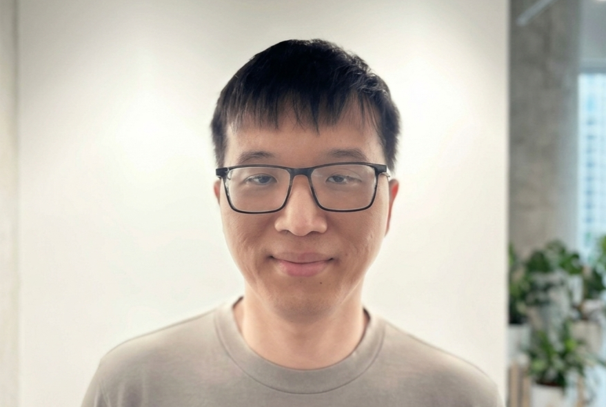 Young man with short black hair and dark rectangular glasses, smiling slightly against a light background.