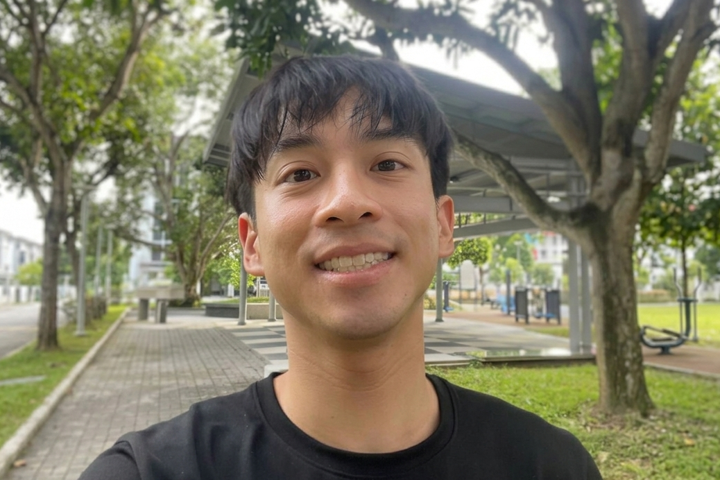 Young man with short dark hair wearing a black shirt smiling outdoors near a covered walkway and trees.
