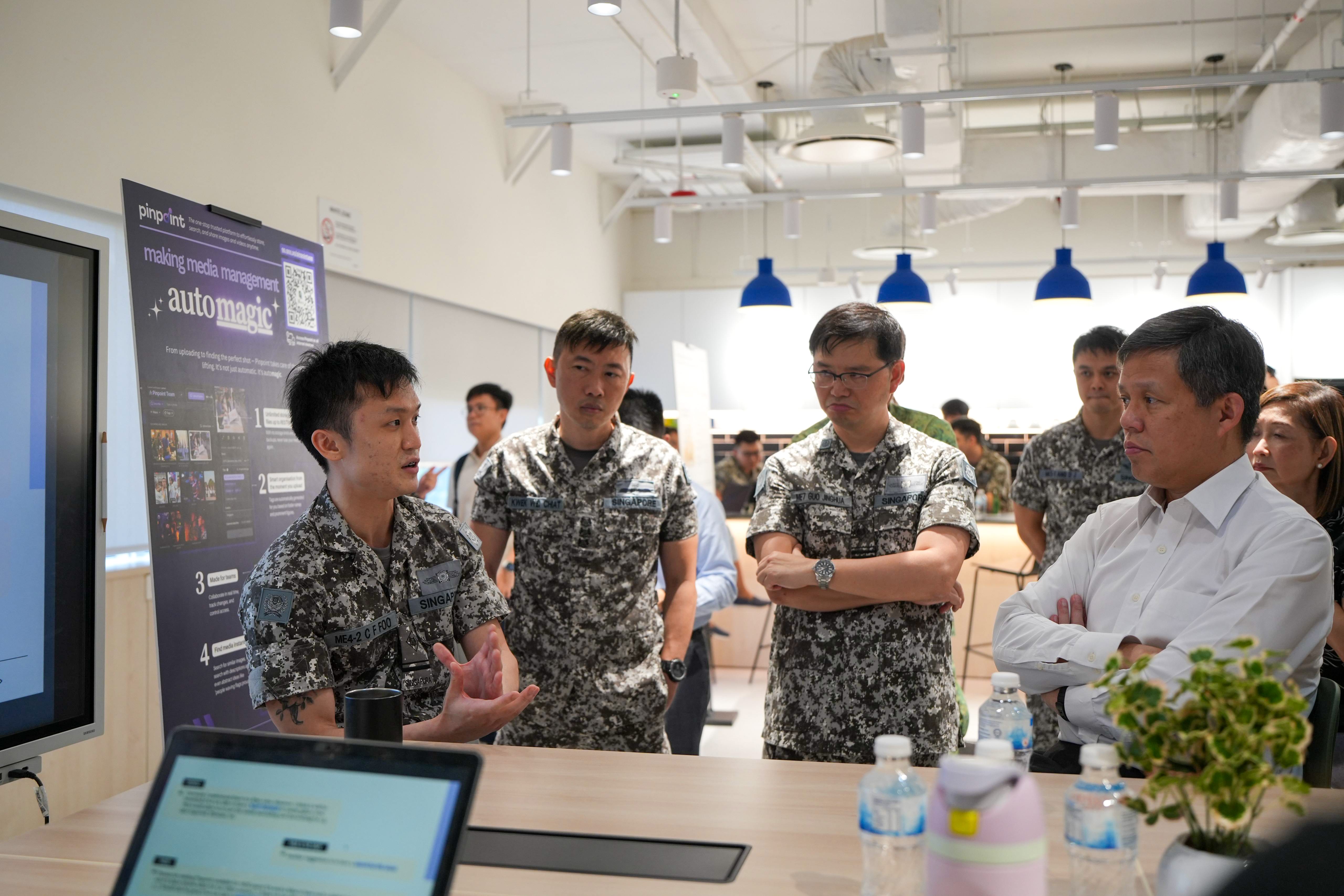 Man in camouflage speaks to three uniformed men and two civilians around a table in a bright office.