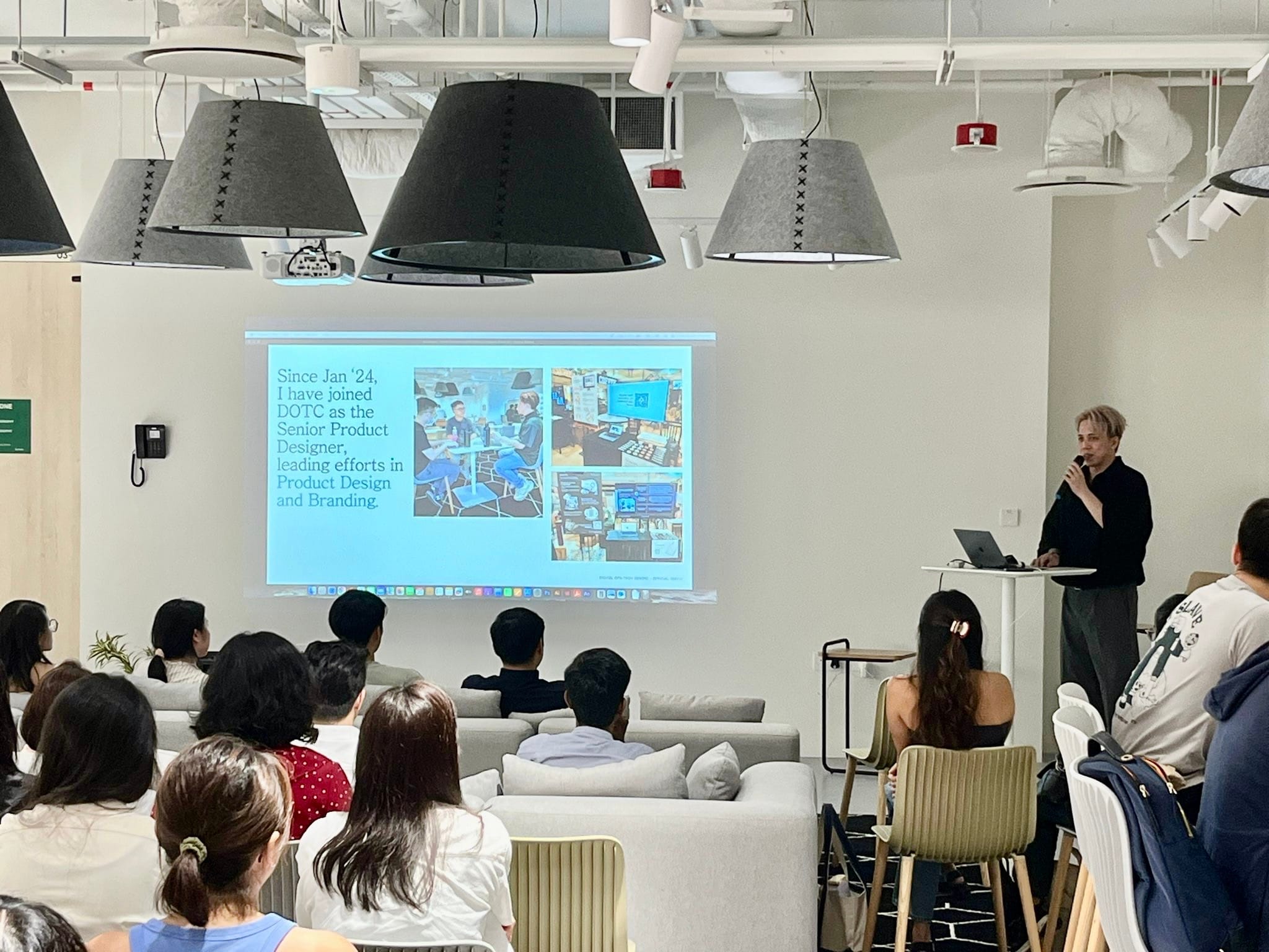 A speaker presents to a seated audience in a modern room, with a slide about joining DOTC as Senior Product Designer.