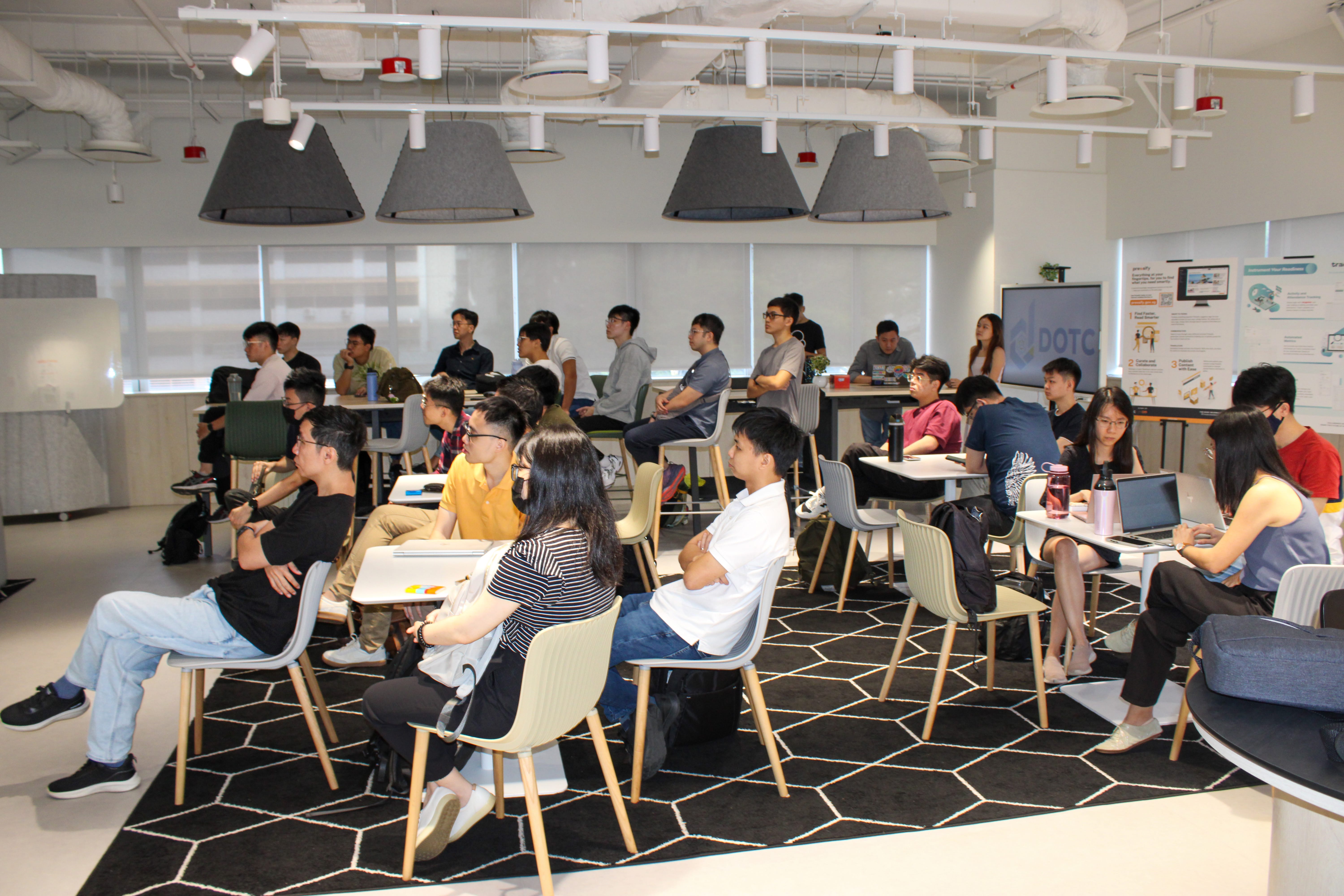 People seated at tables in a bright classroom, listening, with posters and a screen at the front.