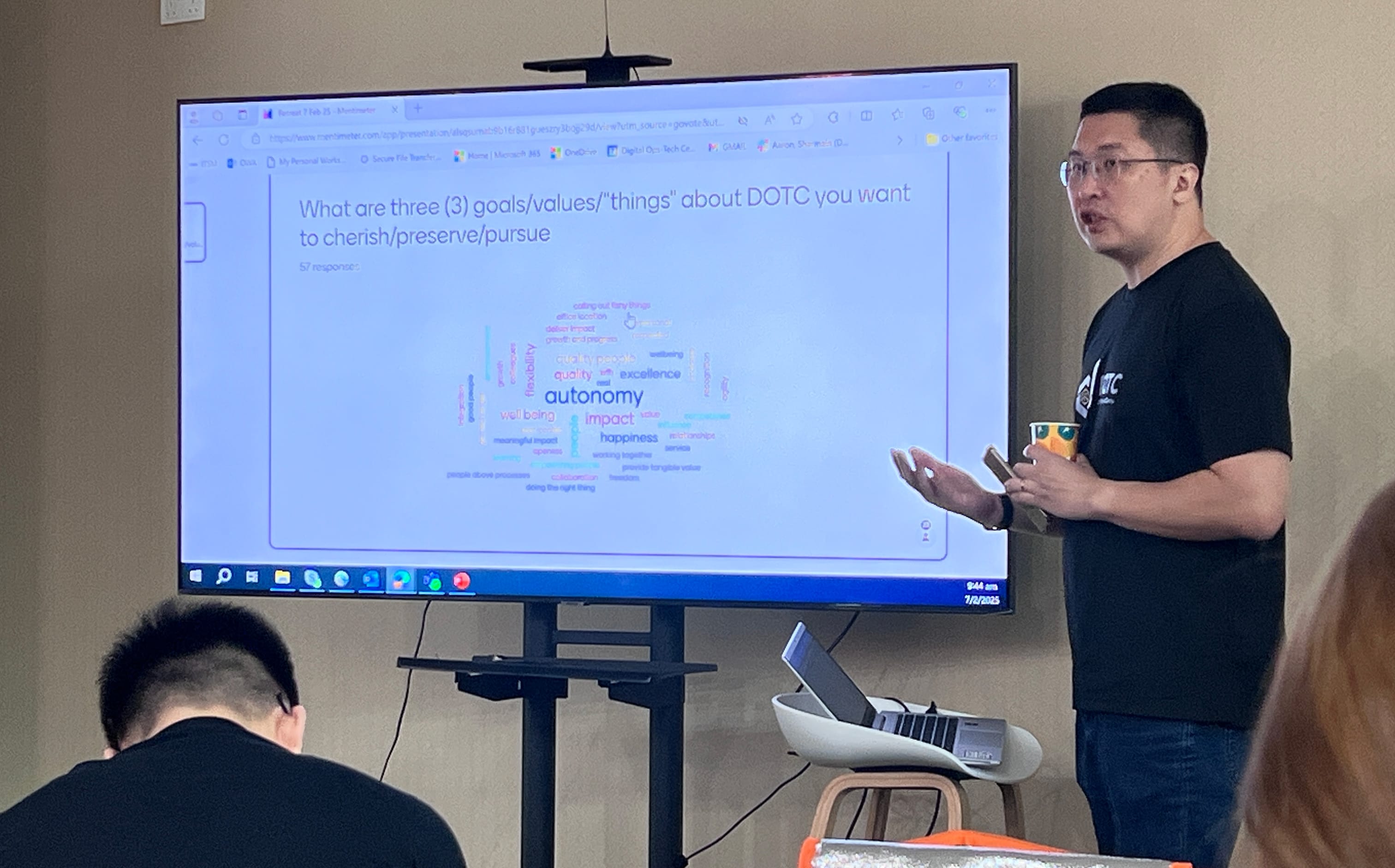 A man in a black shirt presents beside a large screen showing a word cloud about DOTC goals and values.