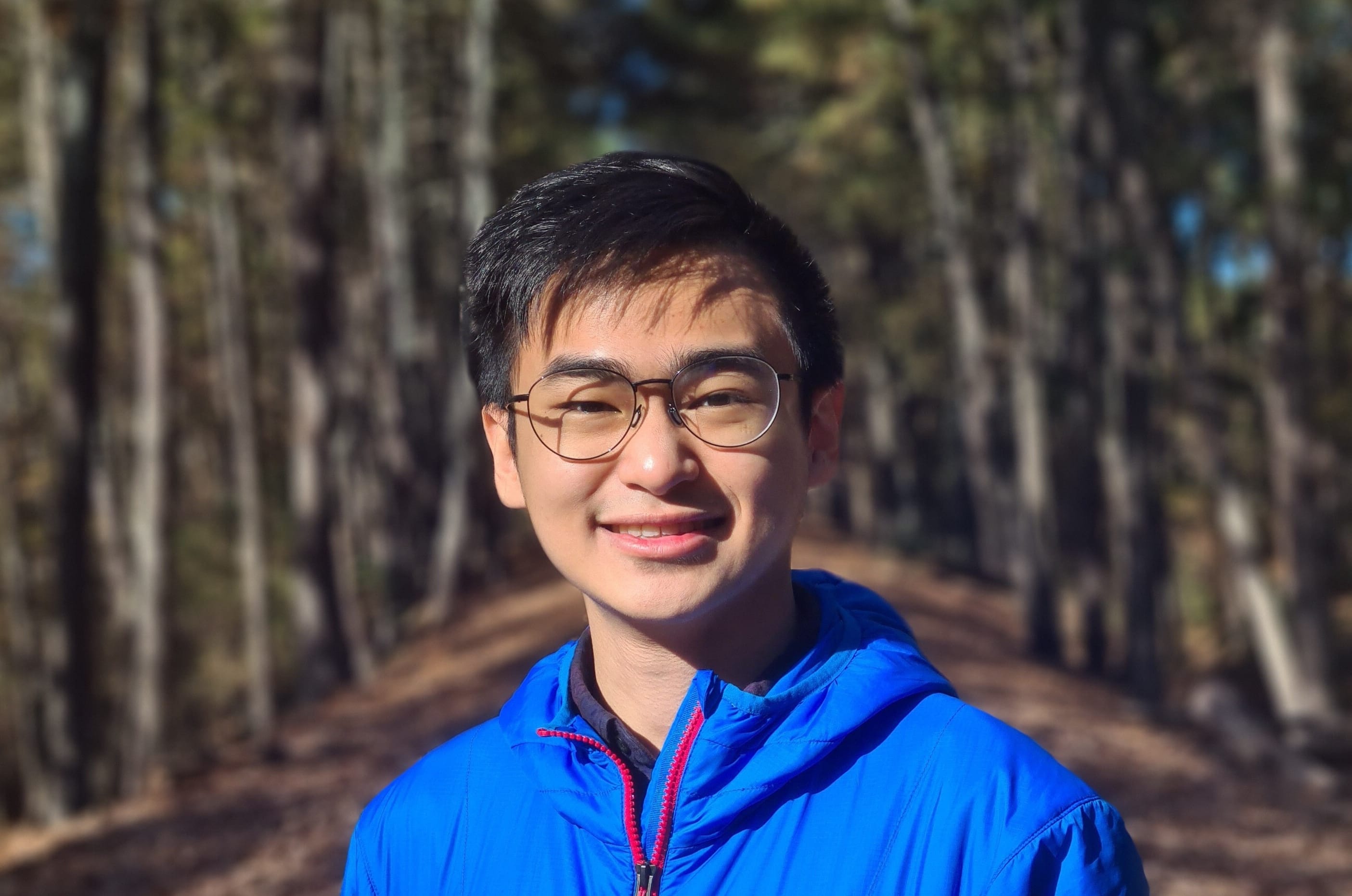 Young man with short dark hair and glasses smiles at the camera, wearing a bright blue jacket on a wooded trail.