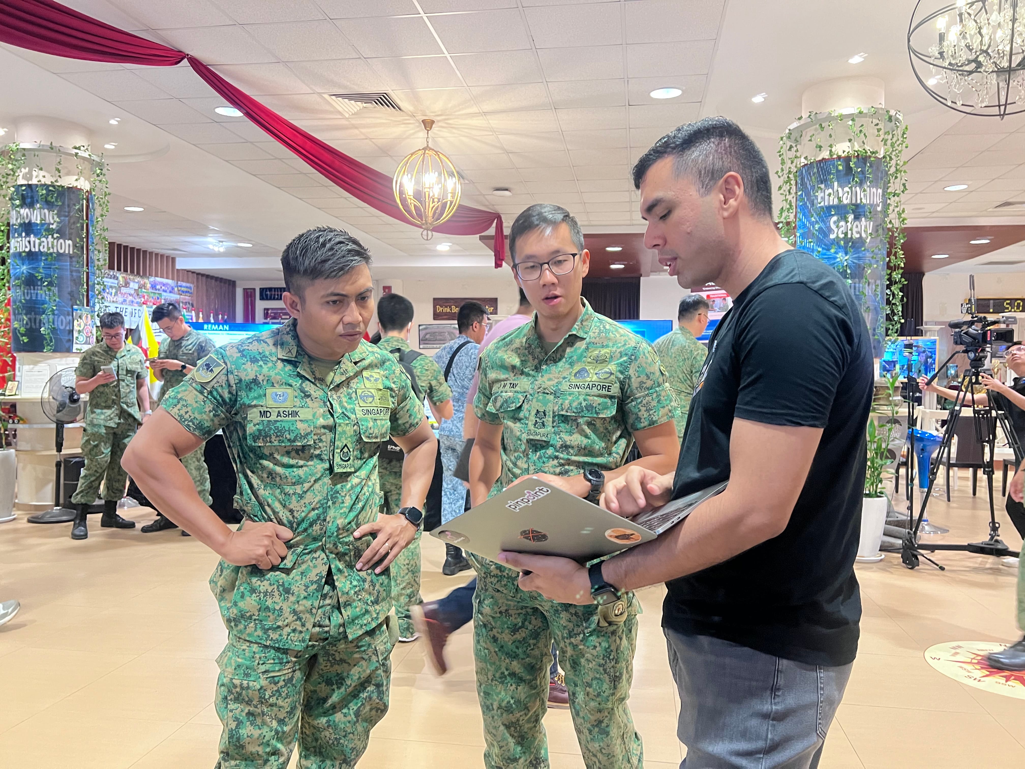 Two Singapore soldiers in camouflage uniforms discuss with a civilian holding a laptop at an indoor event.