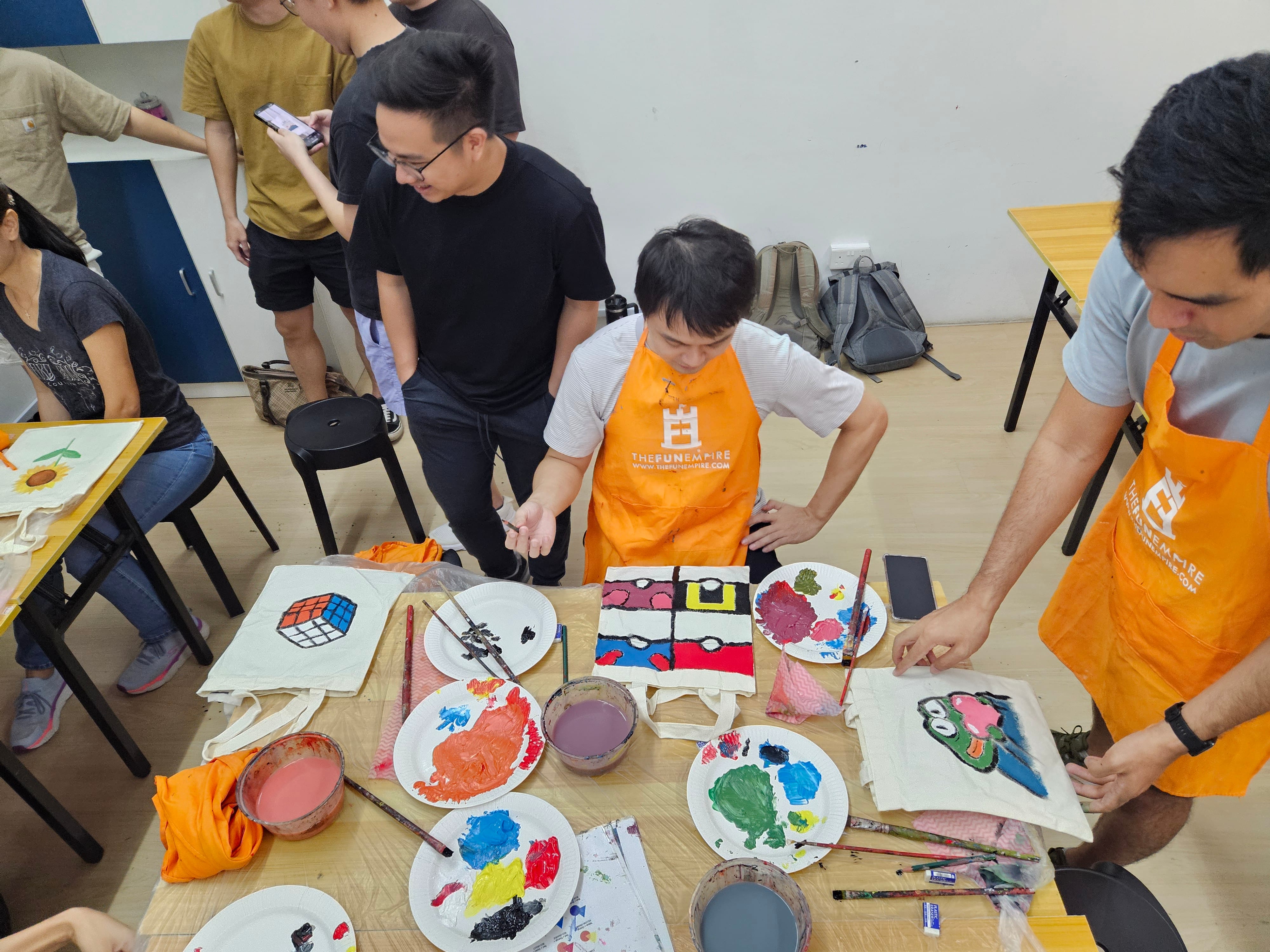 People in orange aprons paint colorful designs on tote bags at a table with paint palettes and brushes.