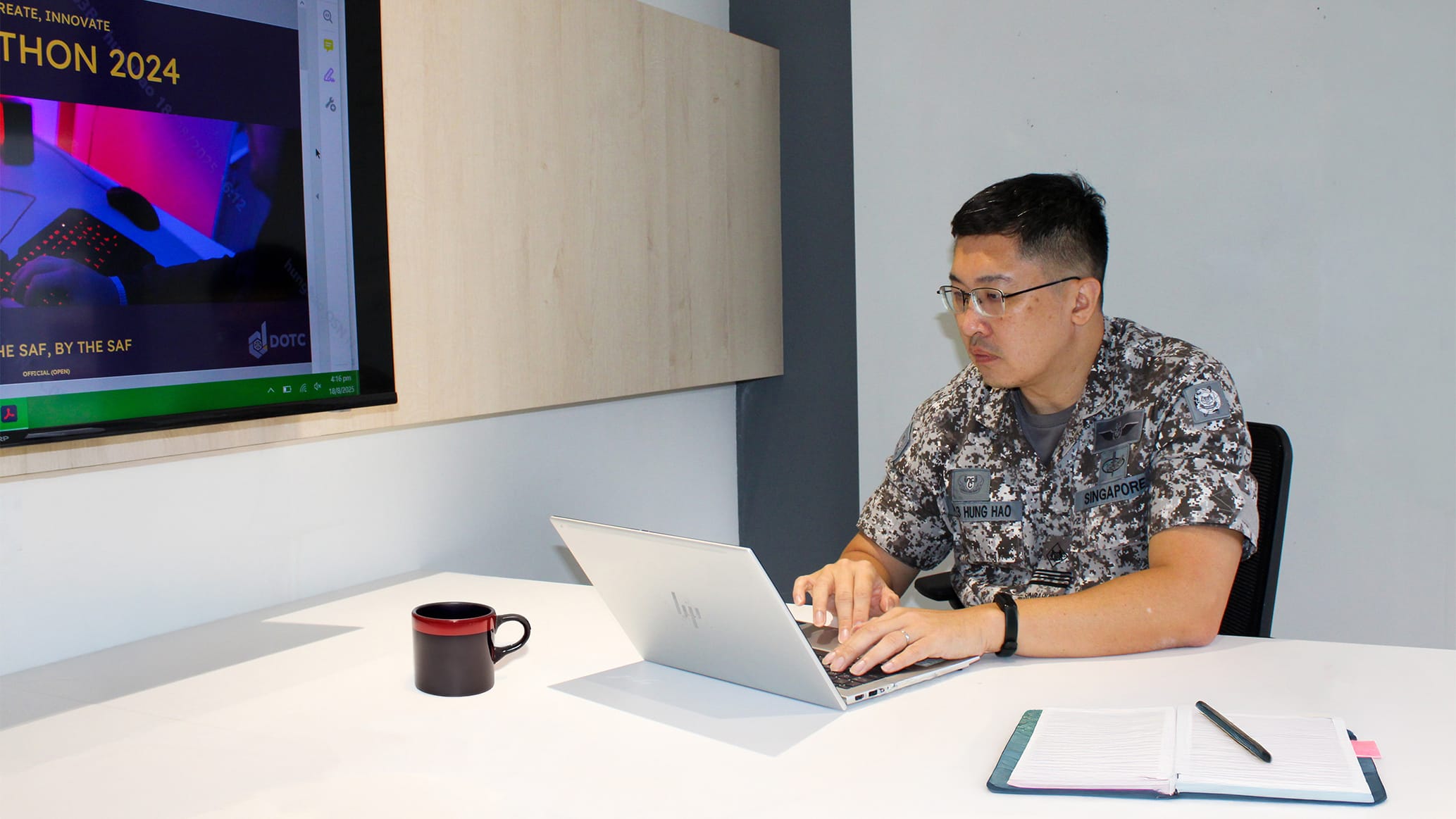 A Singapore military officer in camouflage uniform types on a laptop at a desk with a mug and notebook nearby.