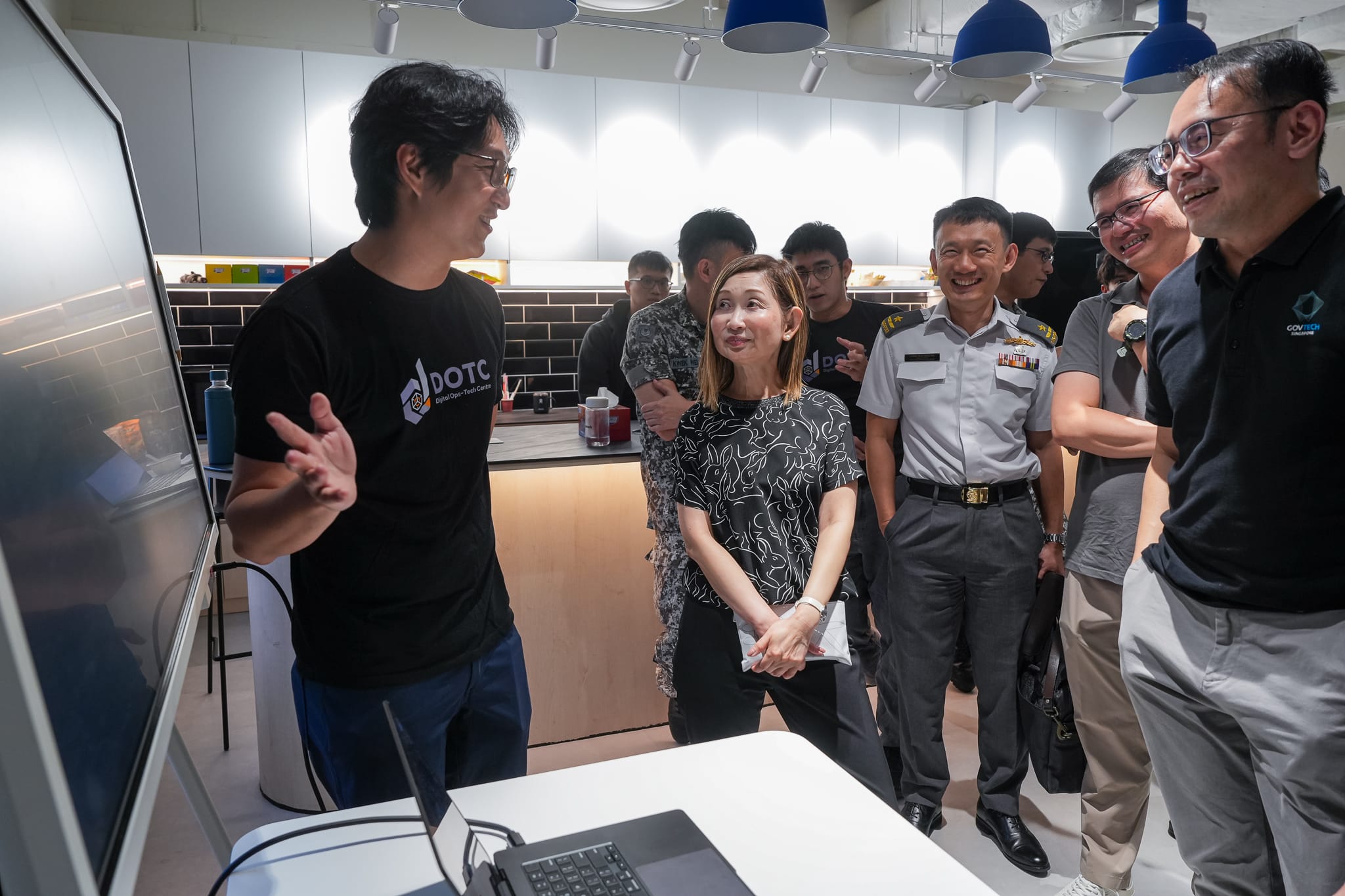 A group of people gathering around a presentation screen indoors, with a man in a black graphic t-shirt gesturing toward the display while others, including a military officer, look on attentively.