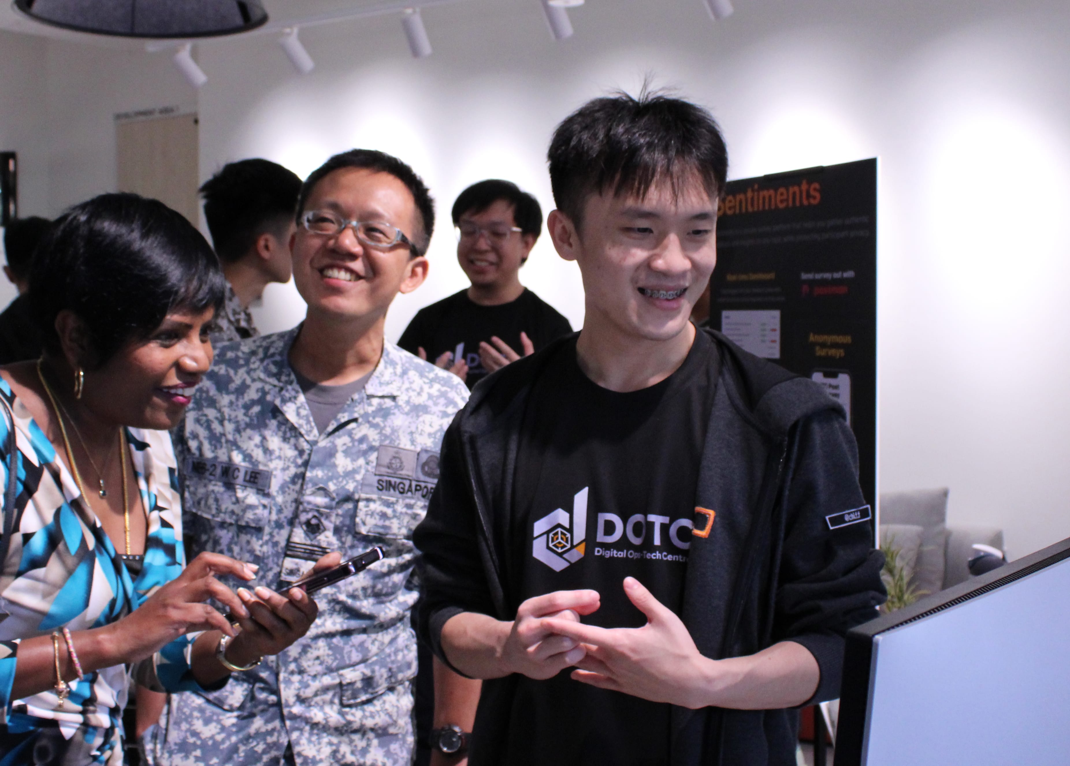 Three people smiling indoors; one in camouflage uniform, one woman holding a phone, one man in black shirt.