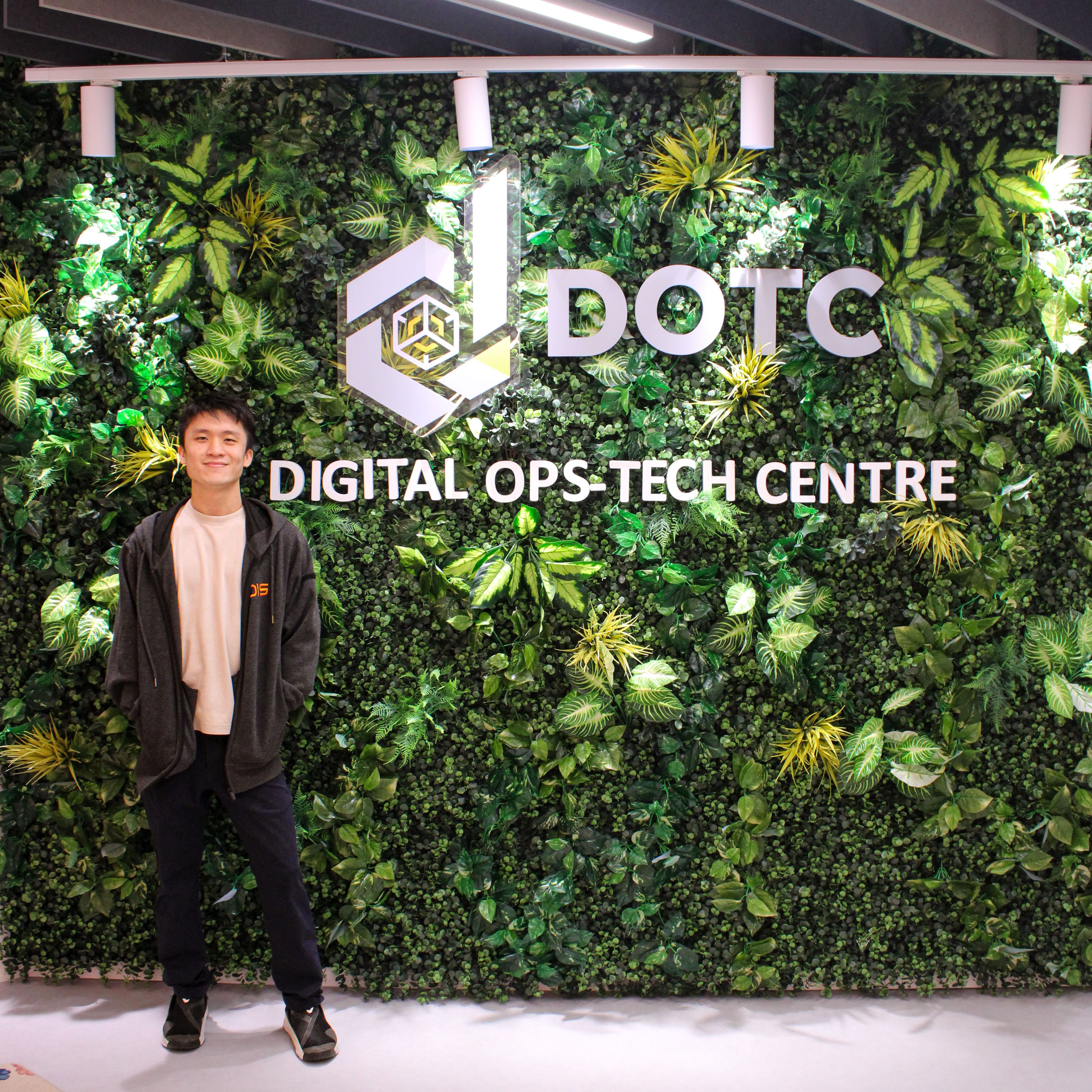 Young man smiling in front of a green plant wall with DOTC Digital Ops-Tech Centre signage.