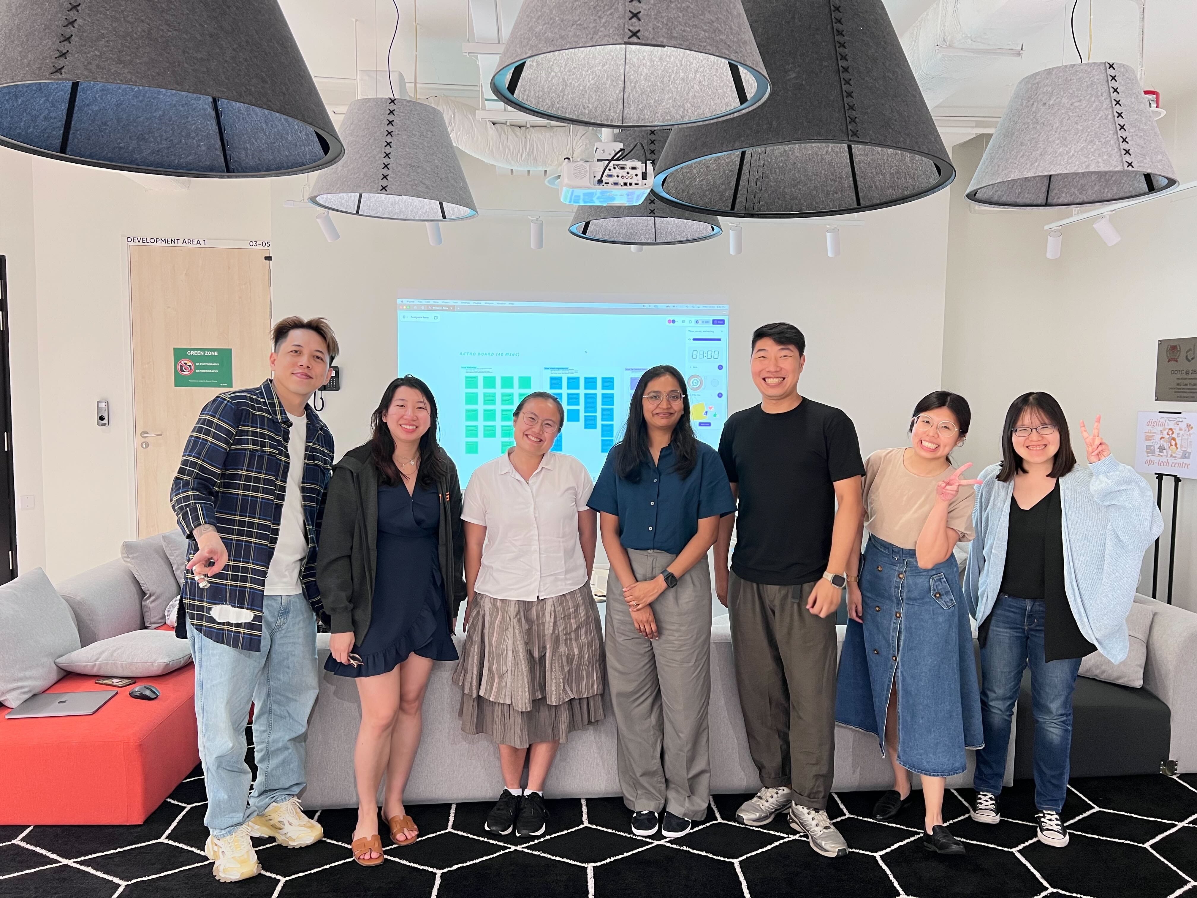 Seven people stand smiling in a modern office lounge with large pendant lamps and a projected screen behind them.