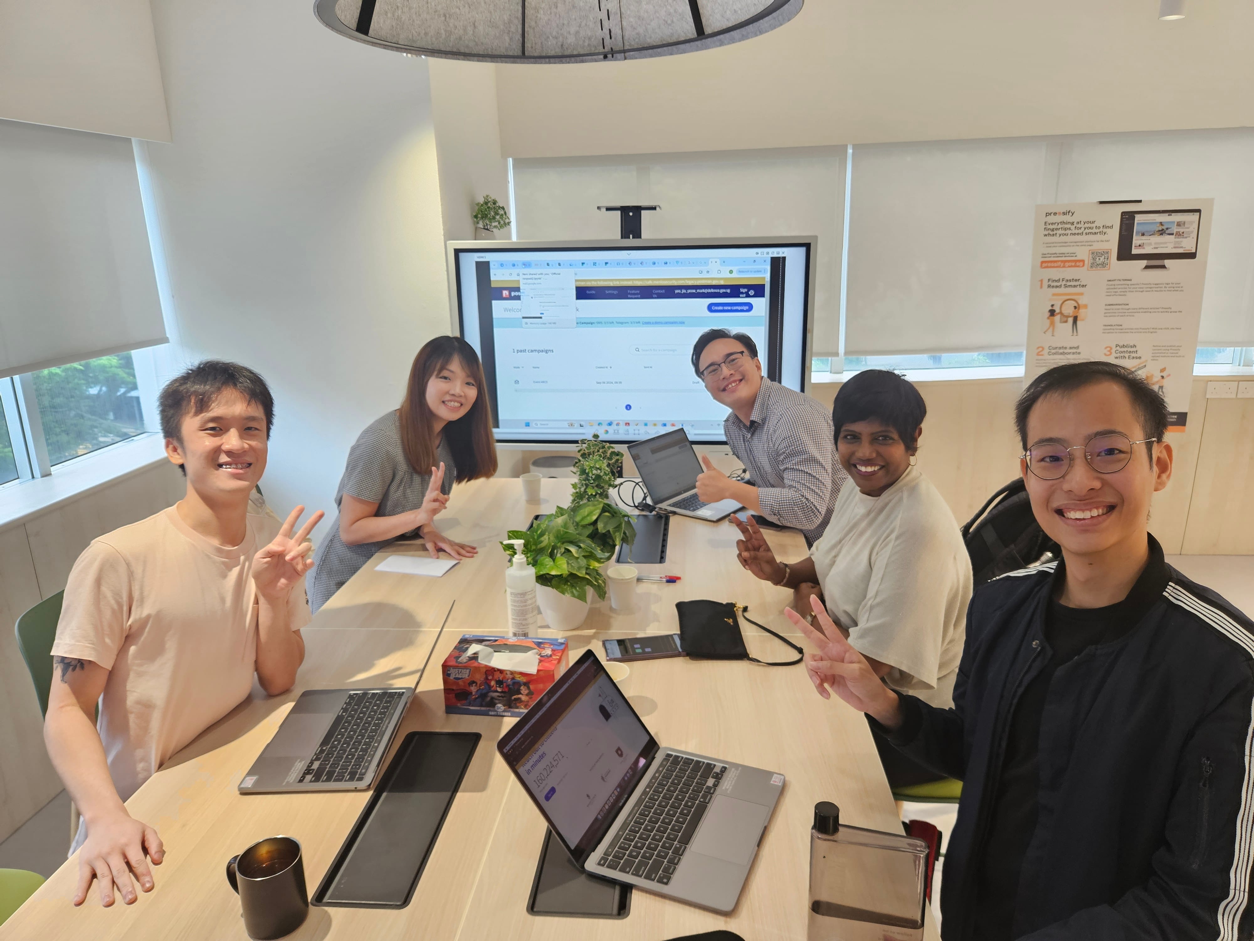 Five people smile around a conference table with laptops, a plant, and a large screen displaying a webpage behind them.
