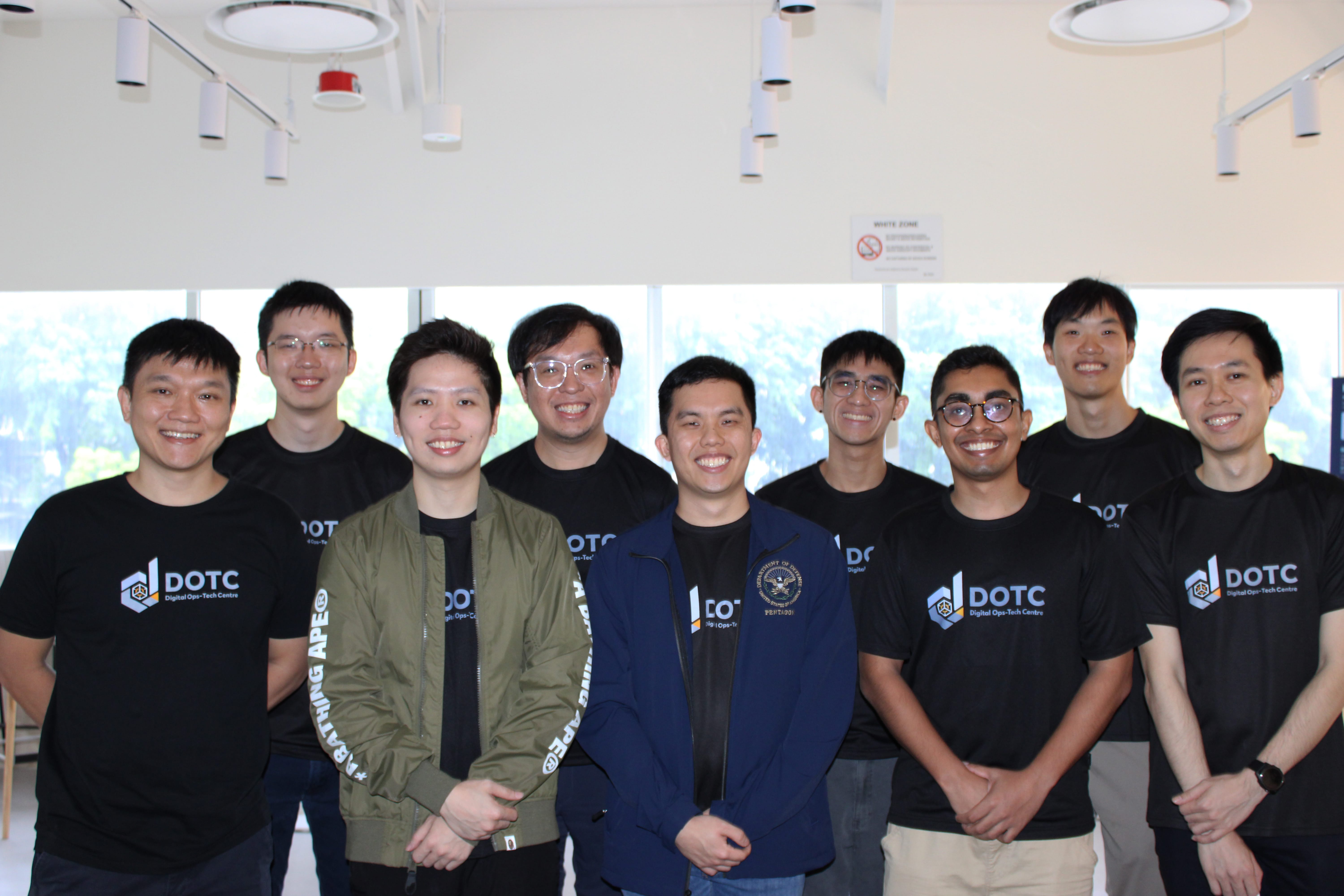 Nine smiling people pose indoors, most wearing black DOTC Digital Ops-Tech Centre t-shirts.