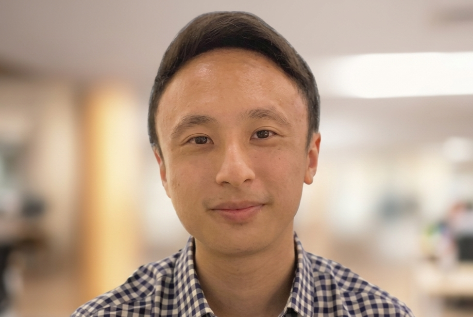 Young Asian man with short dark hair wearing a plaid shirt, smiling softly against a blurred indoor background.