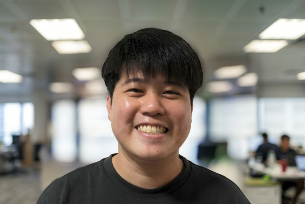 Young man with short black hair wearing a black shirt, smiling broadly in a blurred indoor office setting.
