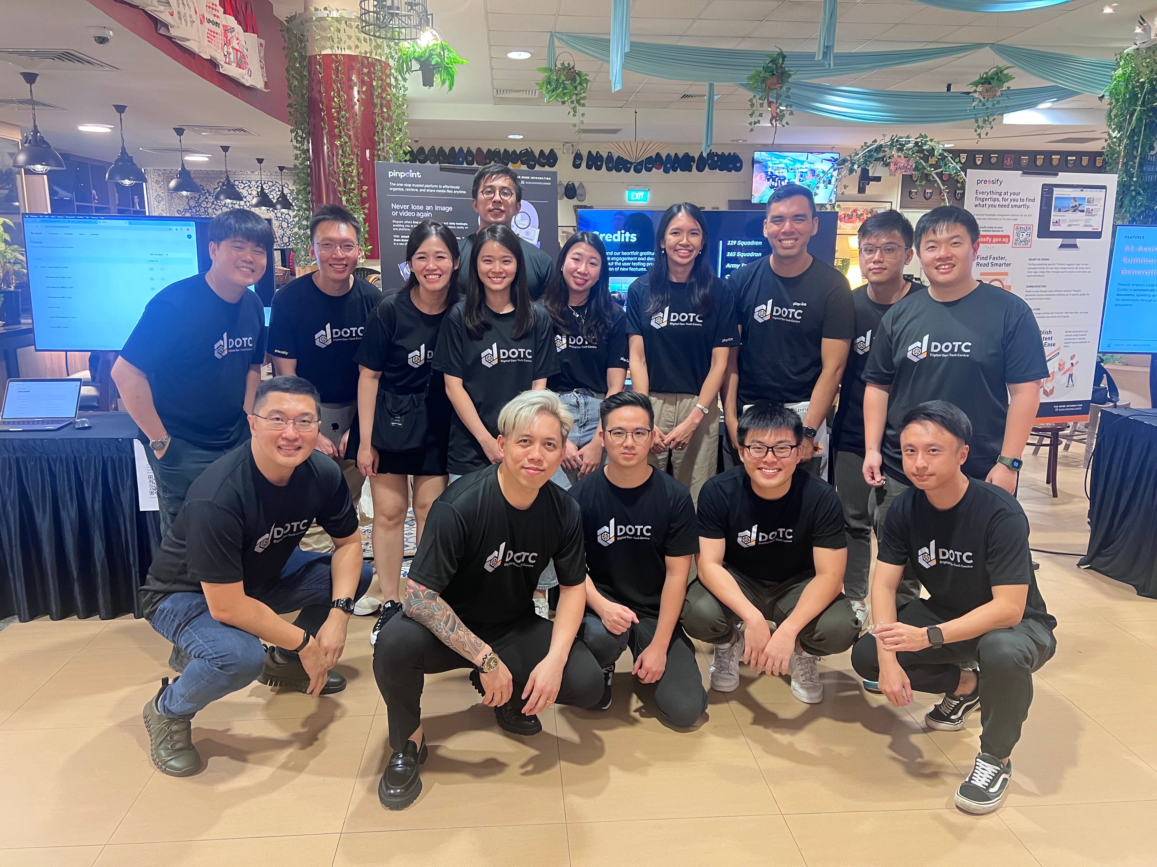 A group of about 15 people wearing matching black DOTC t-shirts pose together indoors at an event.