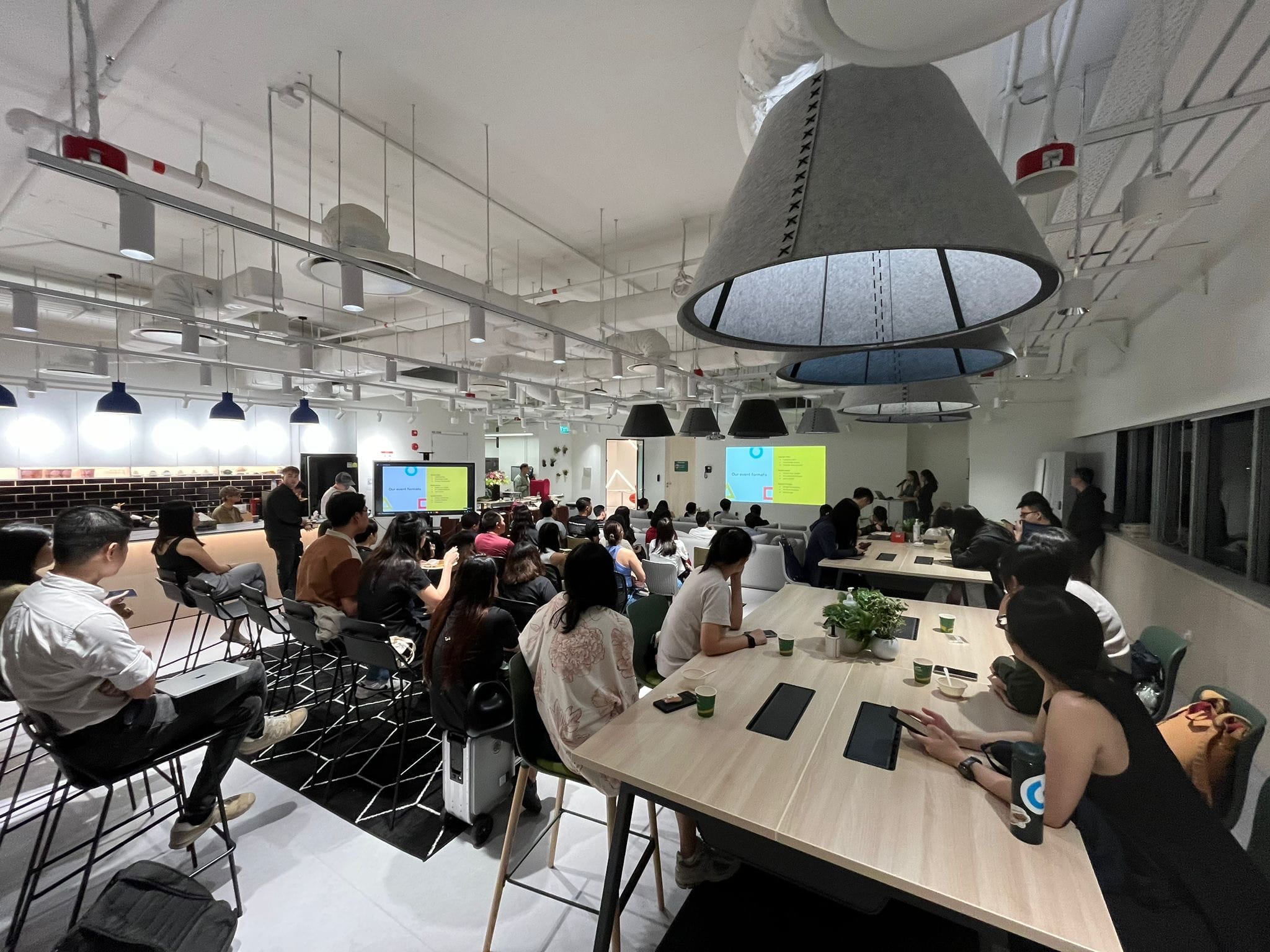 A large group of people seated at tables in a modern co-working space watching two presentation screens.