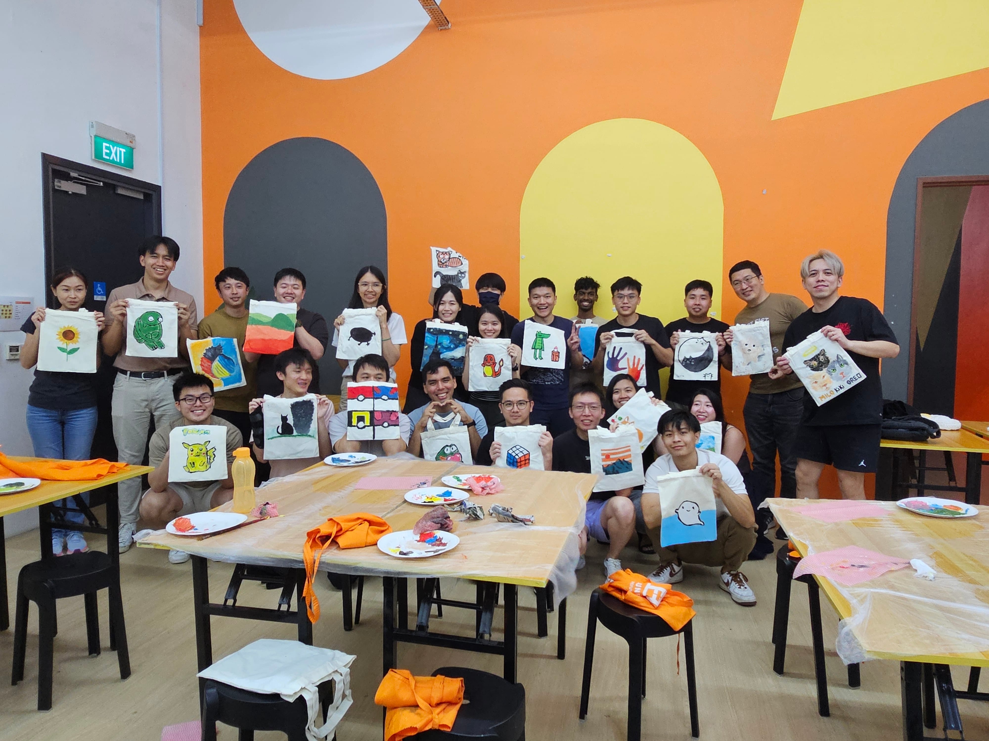 A large group of smiling people hold up hand-painted tote bags in a colorful orange-walled room.