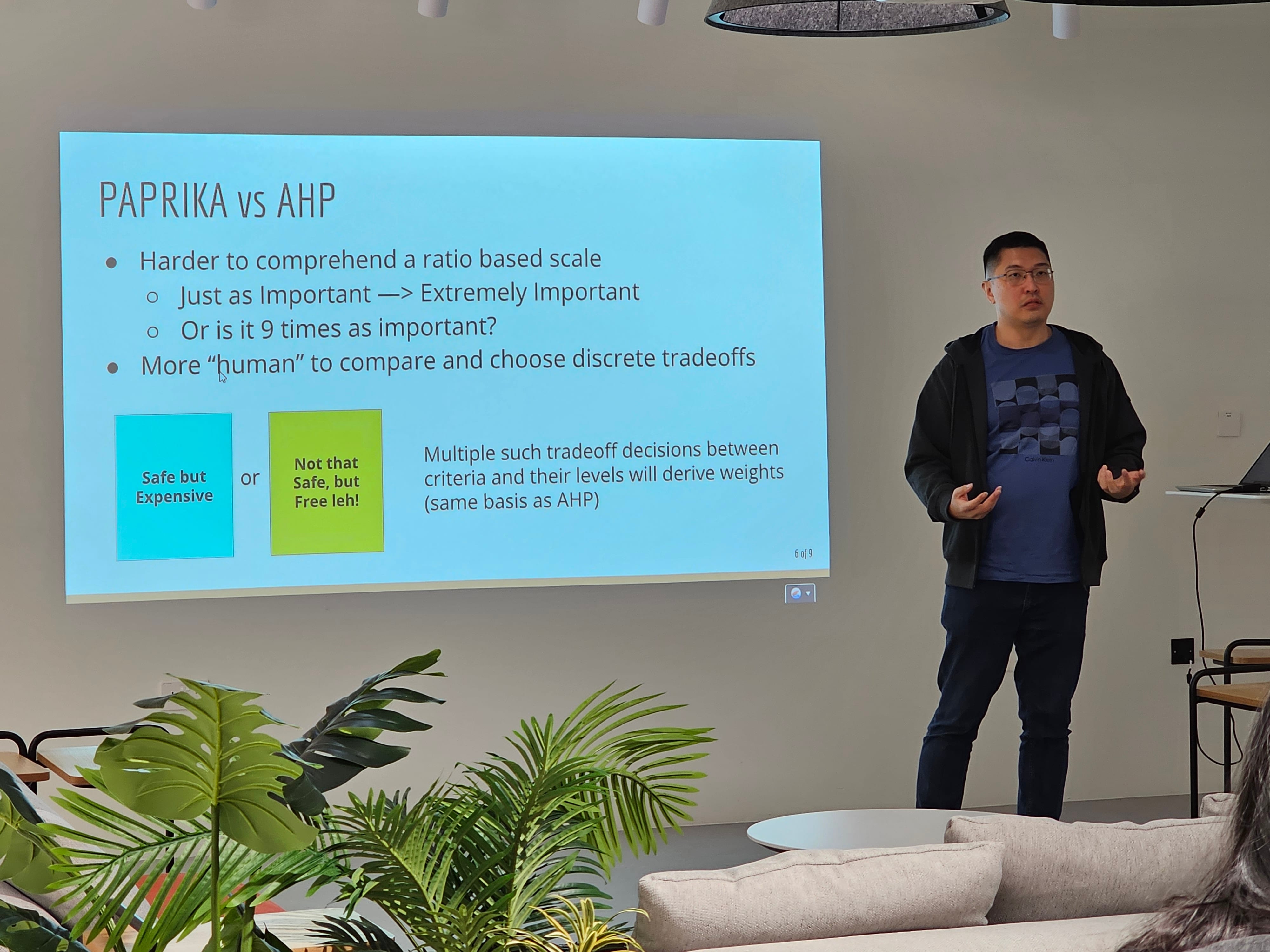 Man presents beside slide titled PAPRIKA vs AHP, with plants and a couch in the foreground.