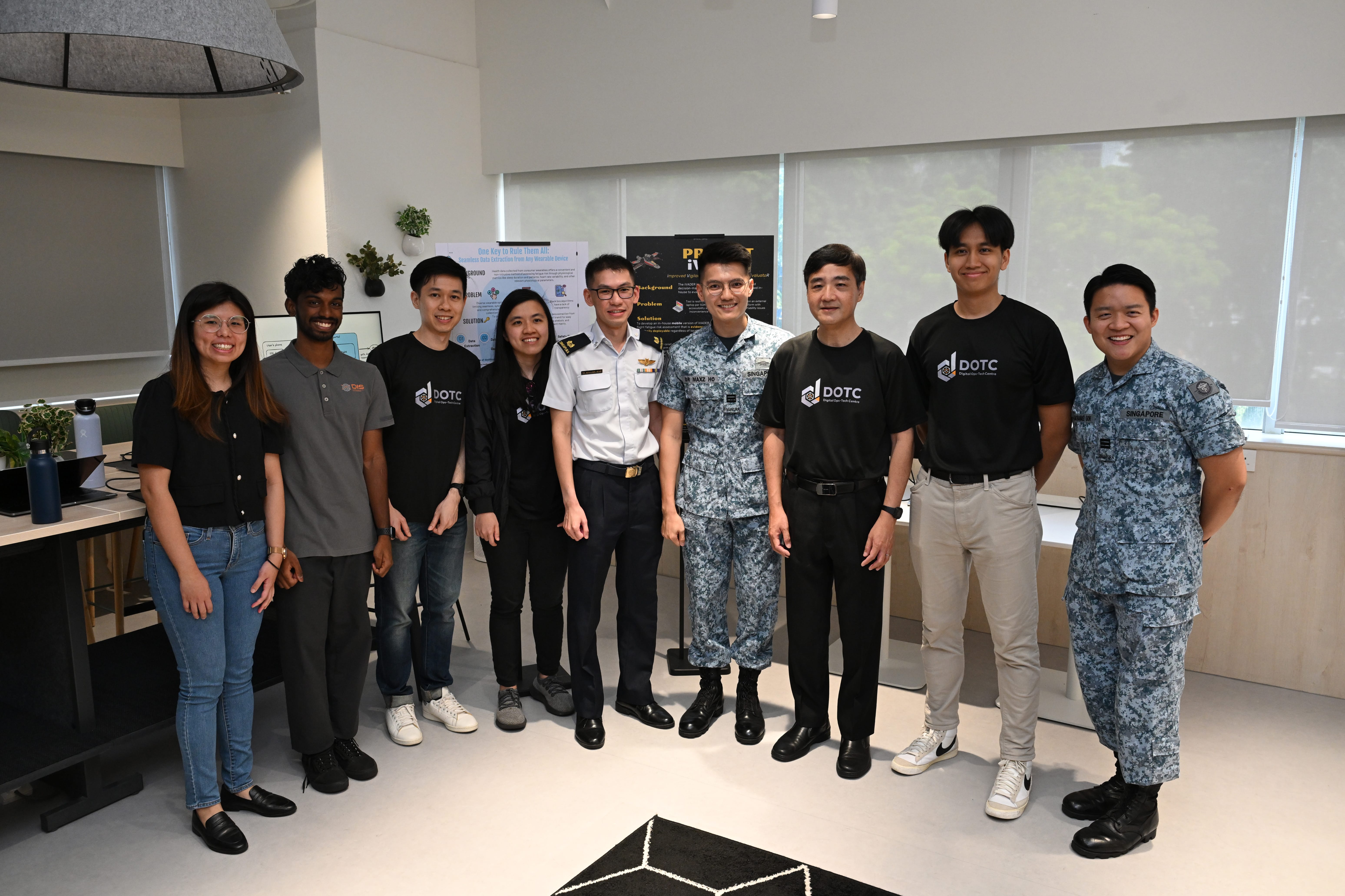 Nine people pose in an office; two in military uniforms and several in black DOTC shirts, all smiling.