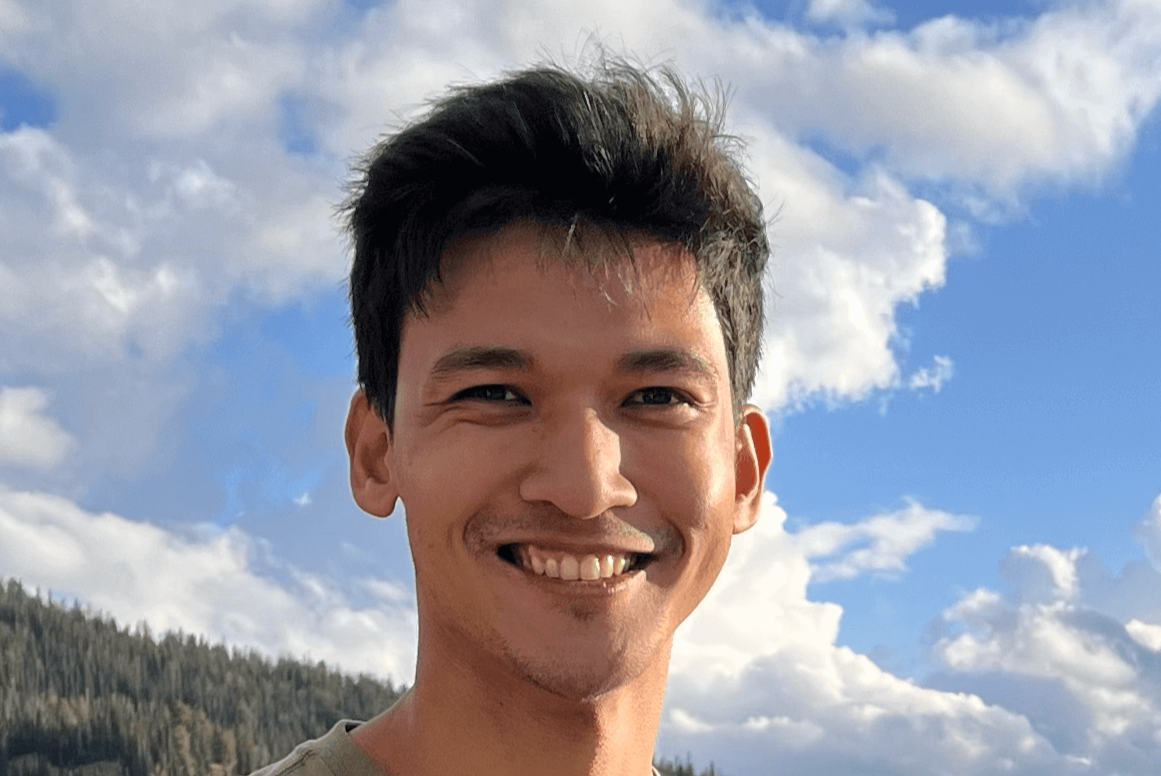 A smiling young man in a tan shirt stands outdoors with forested mountains and a cloudy blue sky behind him.