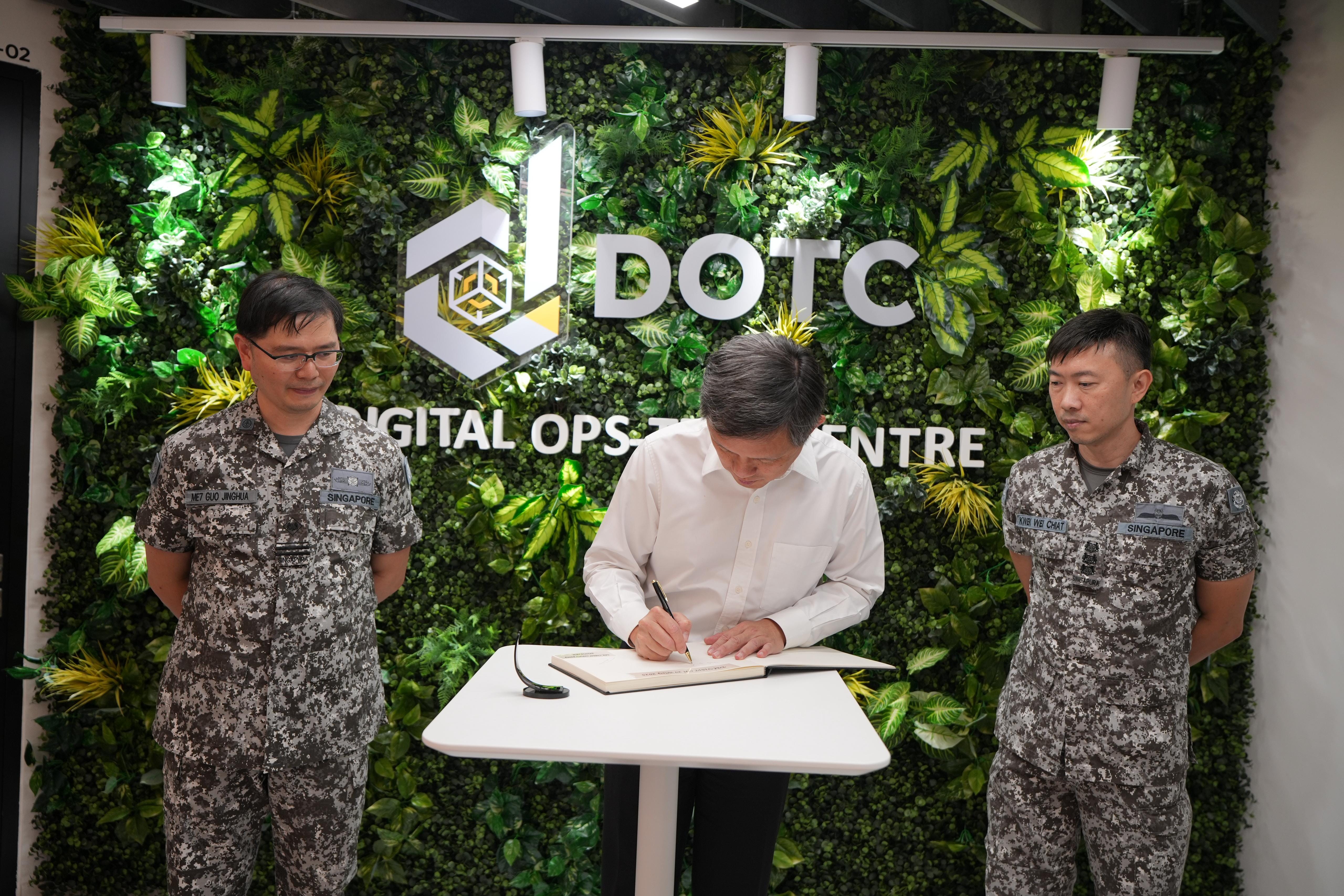 A man in white shirt signs a book at DOTC Digital Ops-Tech Centre, flanked by two Singapore military personnel.