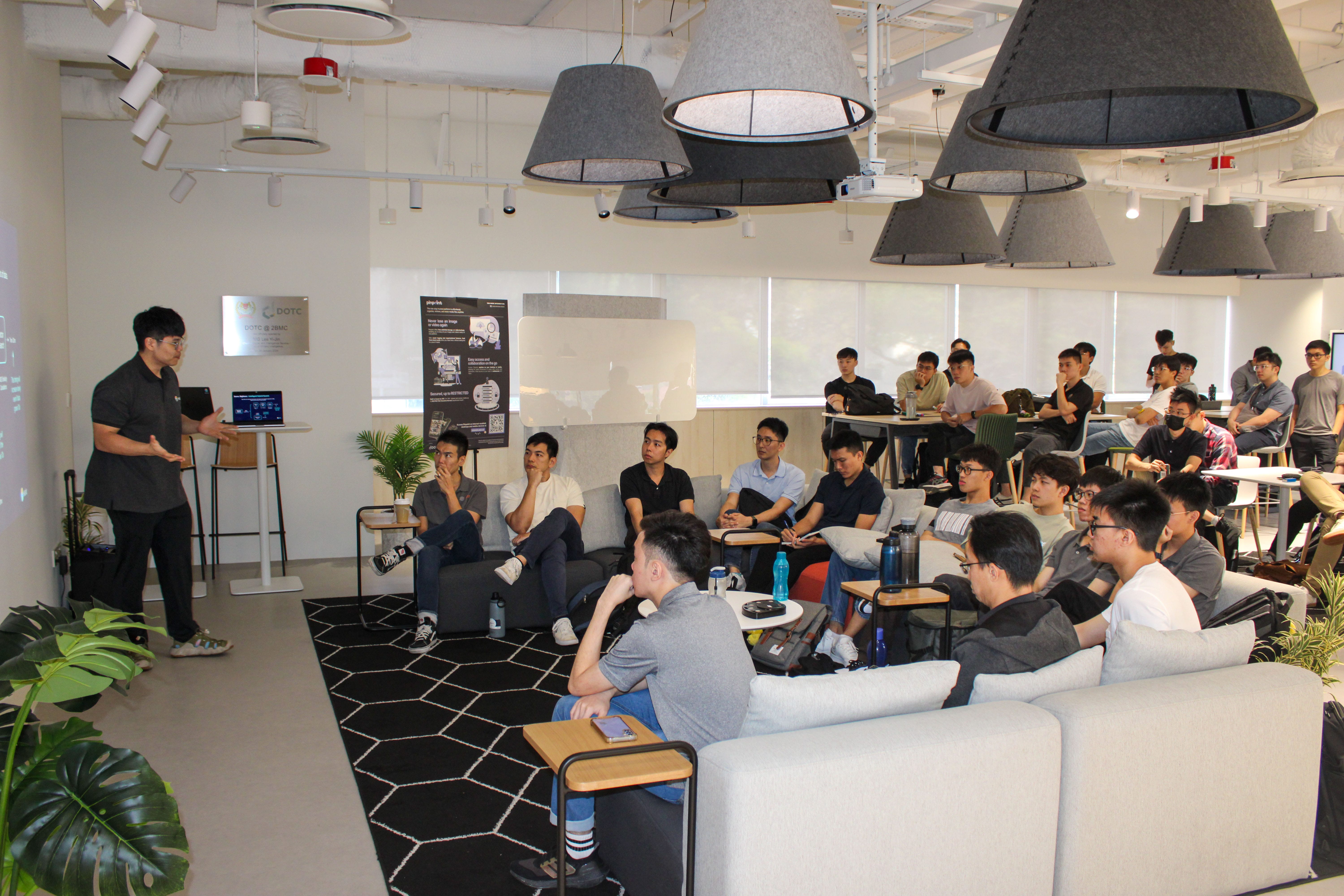 A man presents to a large seated audience in a modern coworking space with large pendant lights overhead.
