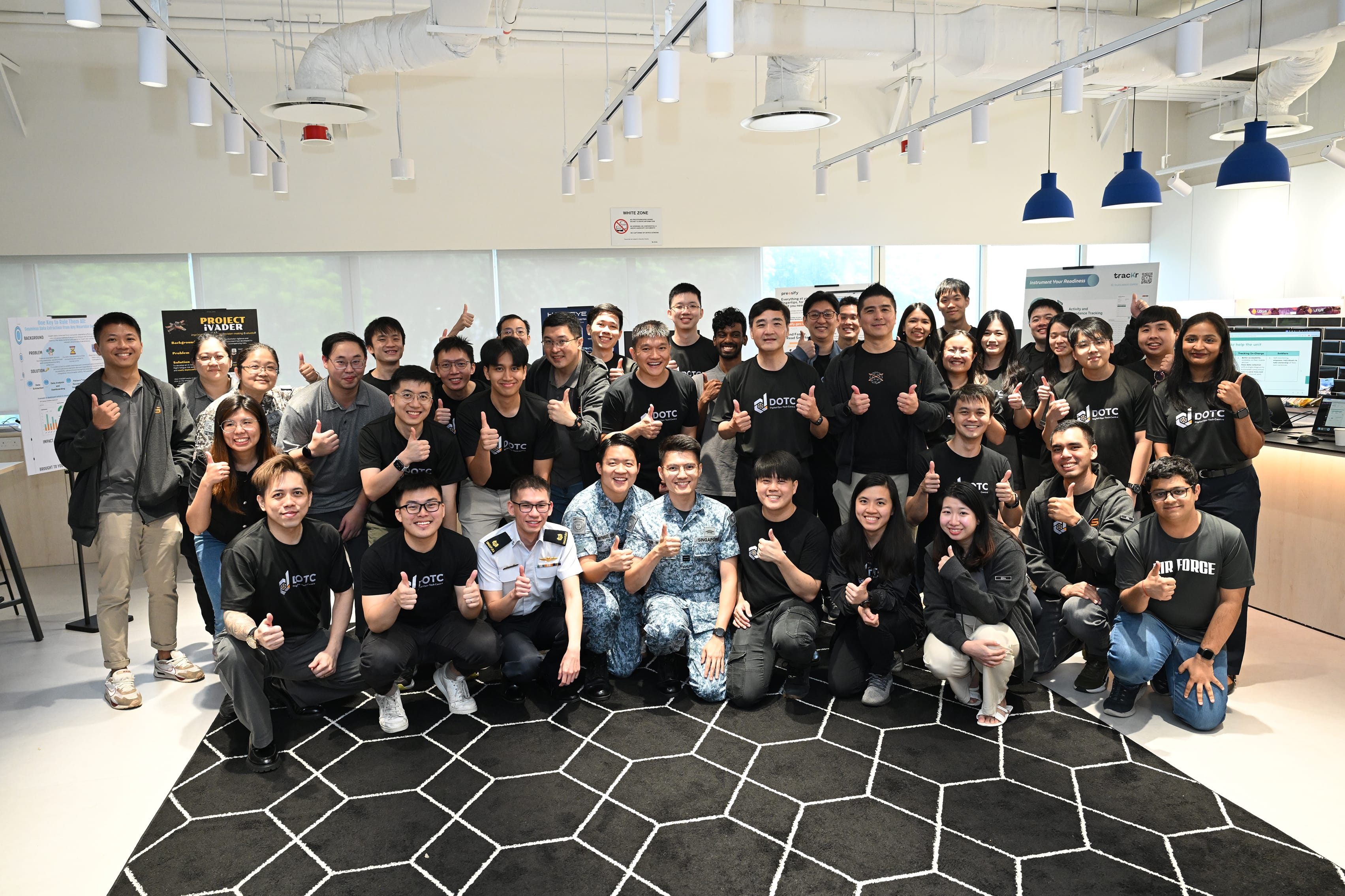 Large group in a bright office, many in black shirts, posing with thumbs up on a patterned rug.