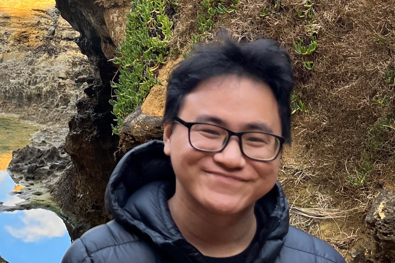Young man with glasses and black puffer jacket smiling in front of rocky coastal cliffs with small plants.
