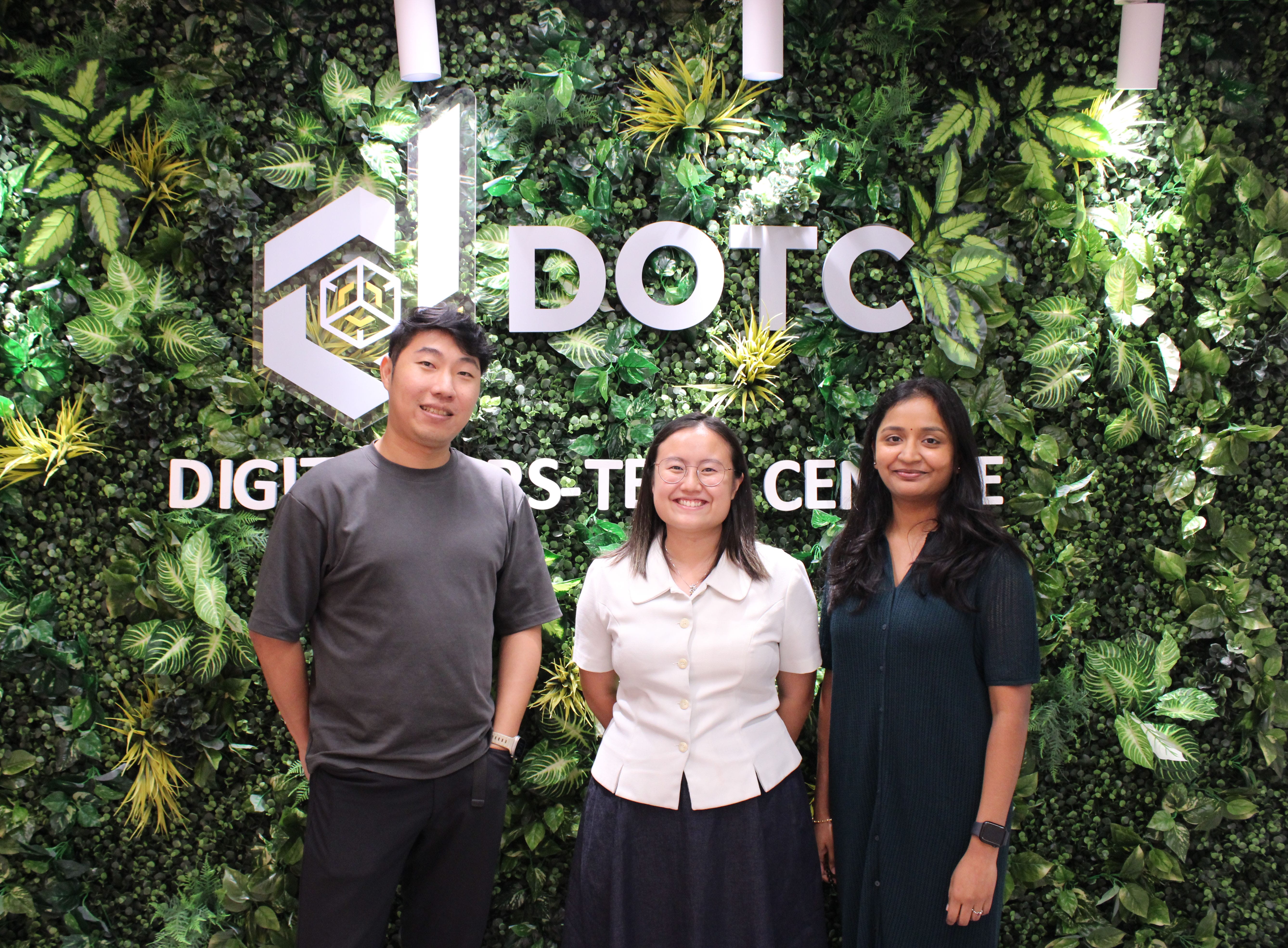 Three smiling people stand in front of a green plant wall displaying the DOTC Digital Cross-Tech Centre sign.