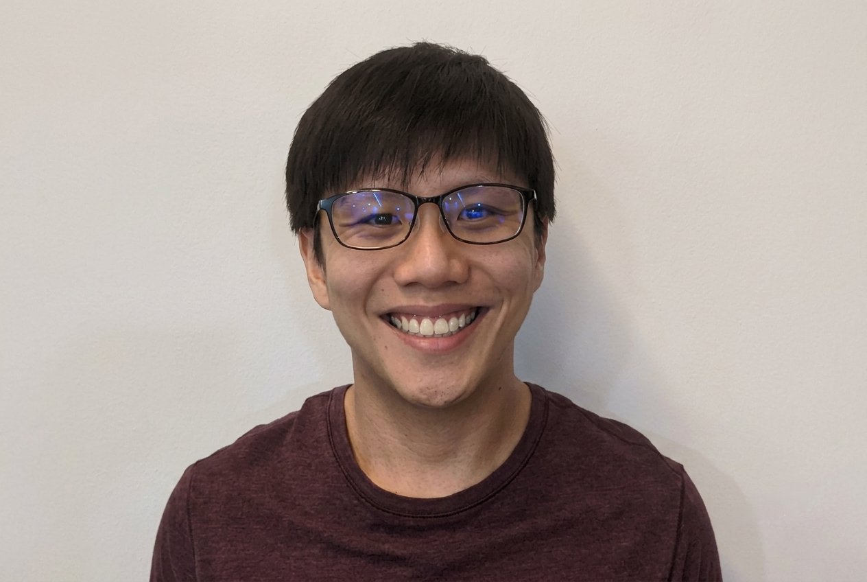 A close-up portrait of a smiling East Asian man with dark hair and rectangular glasses, wearing a dark maroon t-shirt, set against a plain, light-colored wall.
