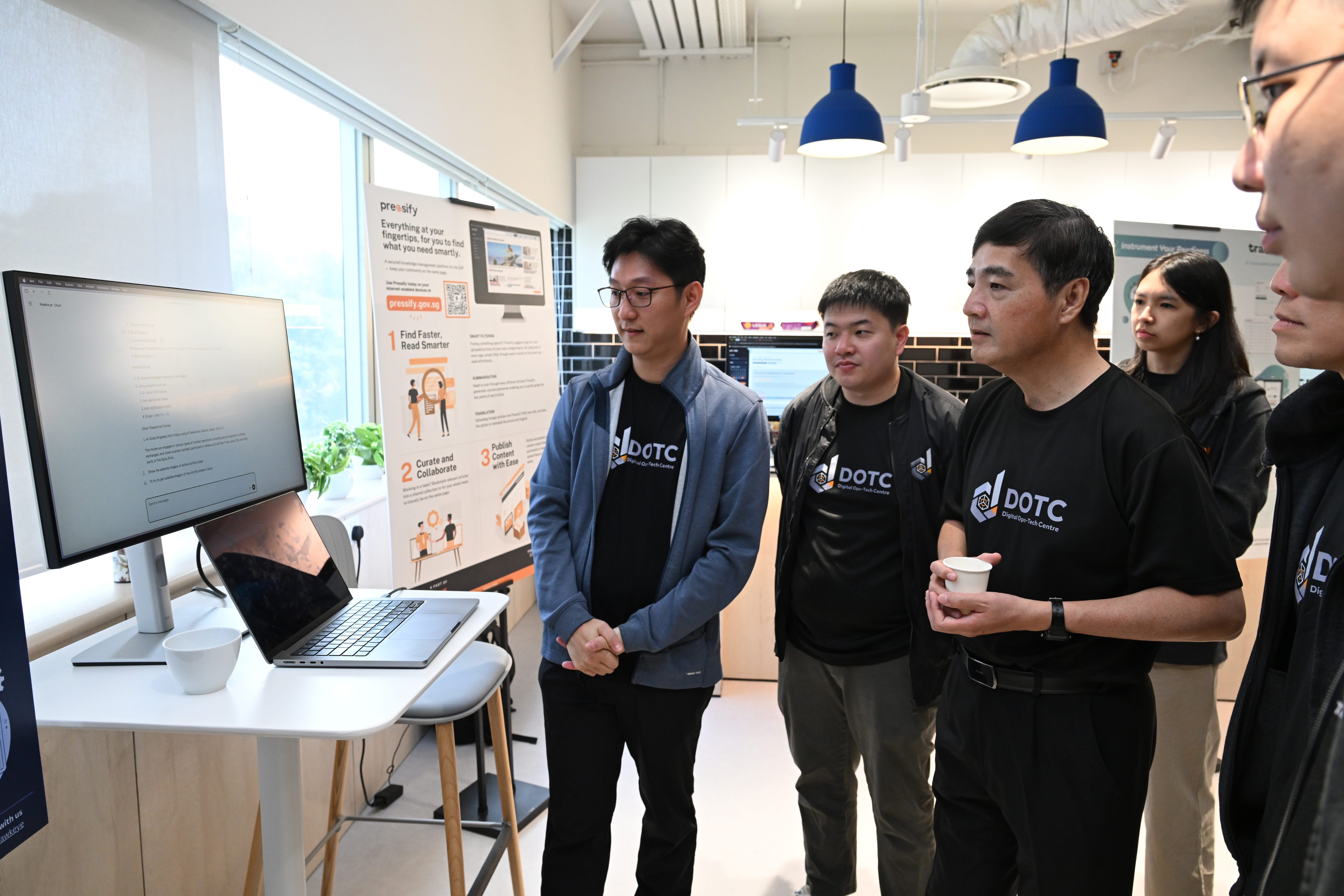 Four people wearing DOTC shirts stand near a desk with a monitor and laptop, viewing a Pressify product poster.
