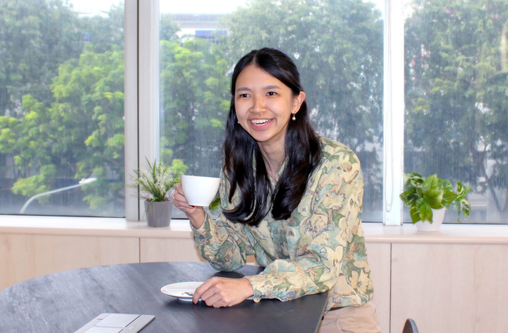 A smiling woman with long dark hair holds a white cup at a table near large windows with green trees outside.
