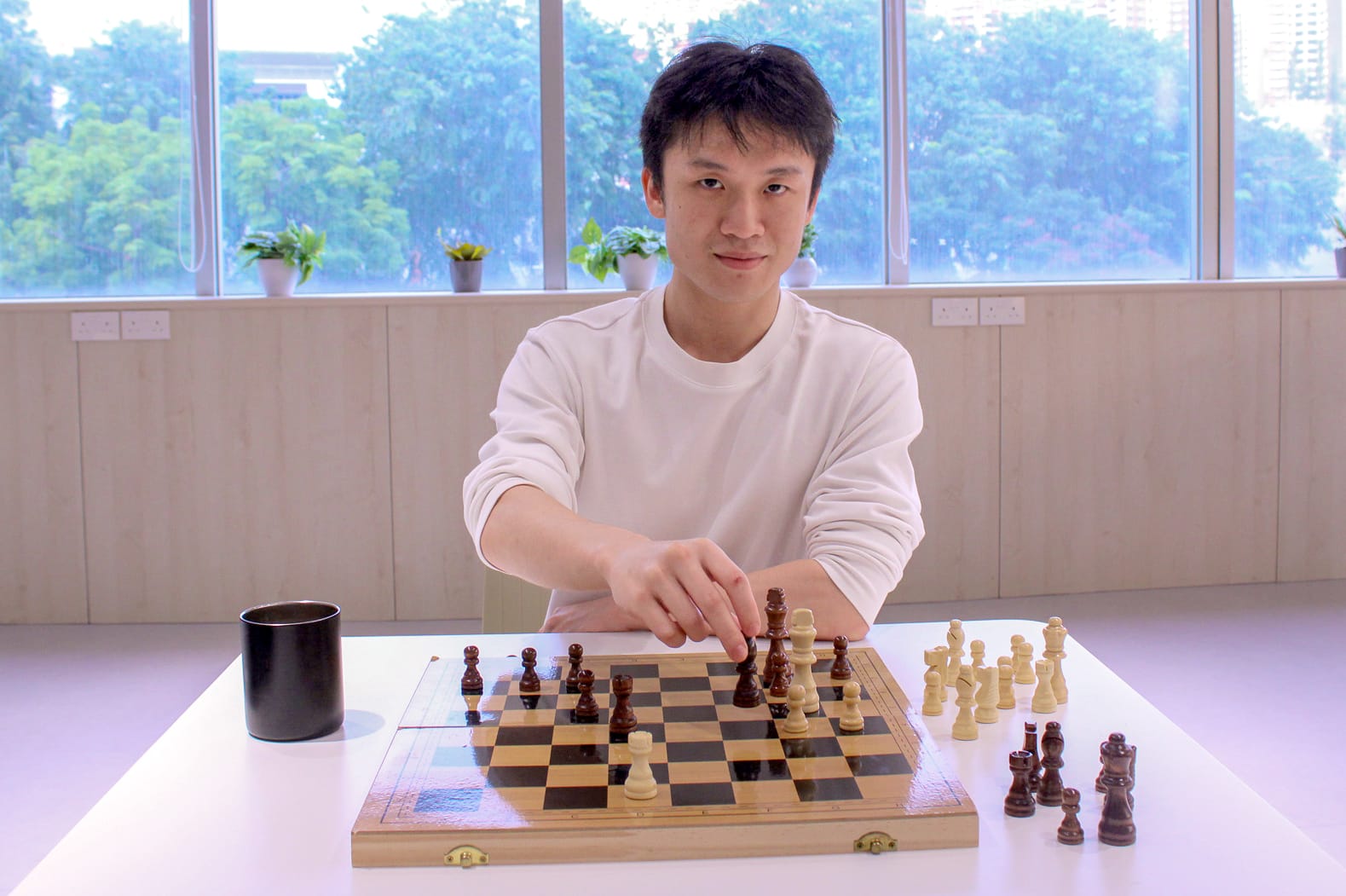 Young man in a white shirt reaching to move a piece on a wooden chess board at a white table.