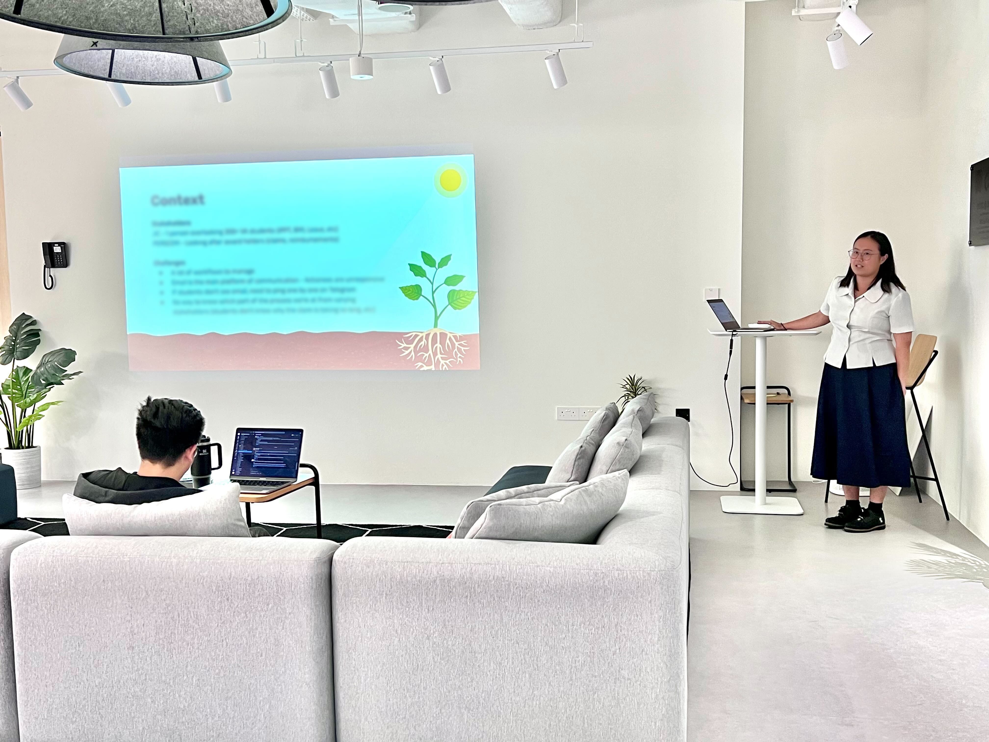 Woman presenting at a standing desk beside a projected slide showing a plant illustration, while a man sits on a sofa with a laptop.