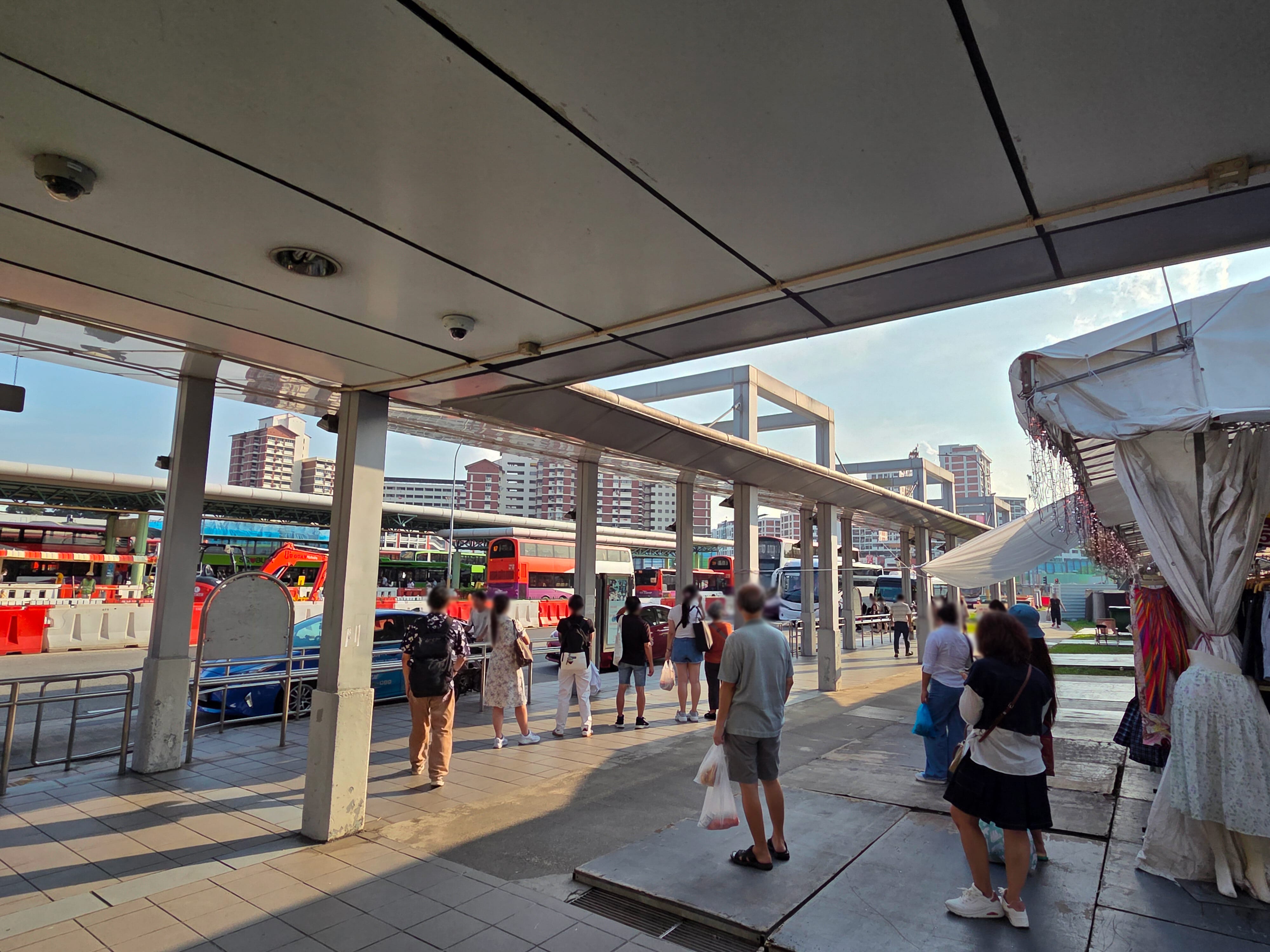 People waiting at a covered bus stop; red buses and tall apartment buildings in the background.