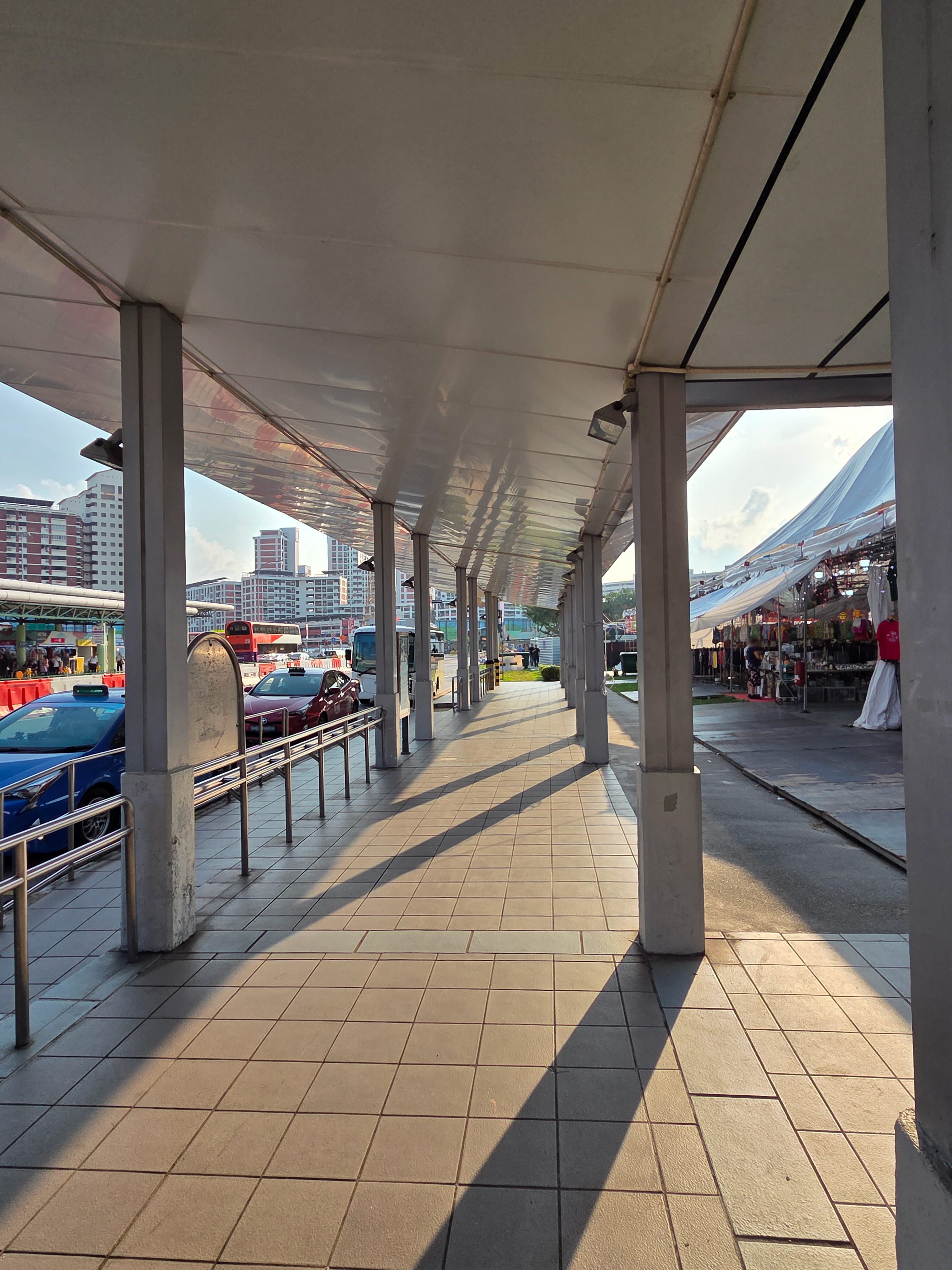 Covered walkway with tiled floor and columns, taxis and buses on left, market tents on right in sun.