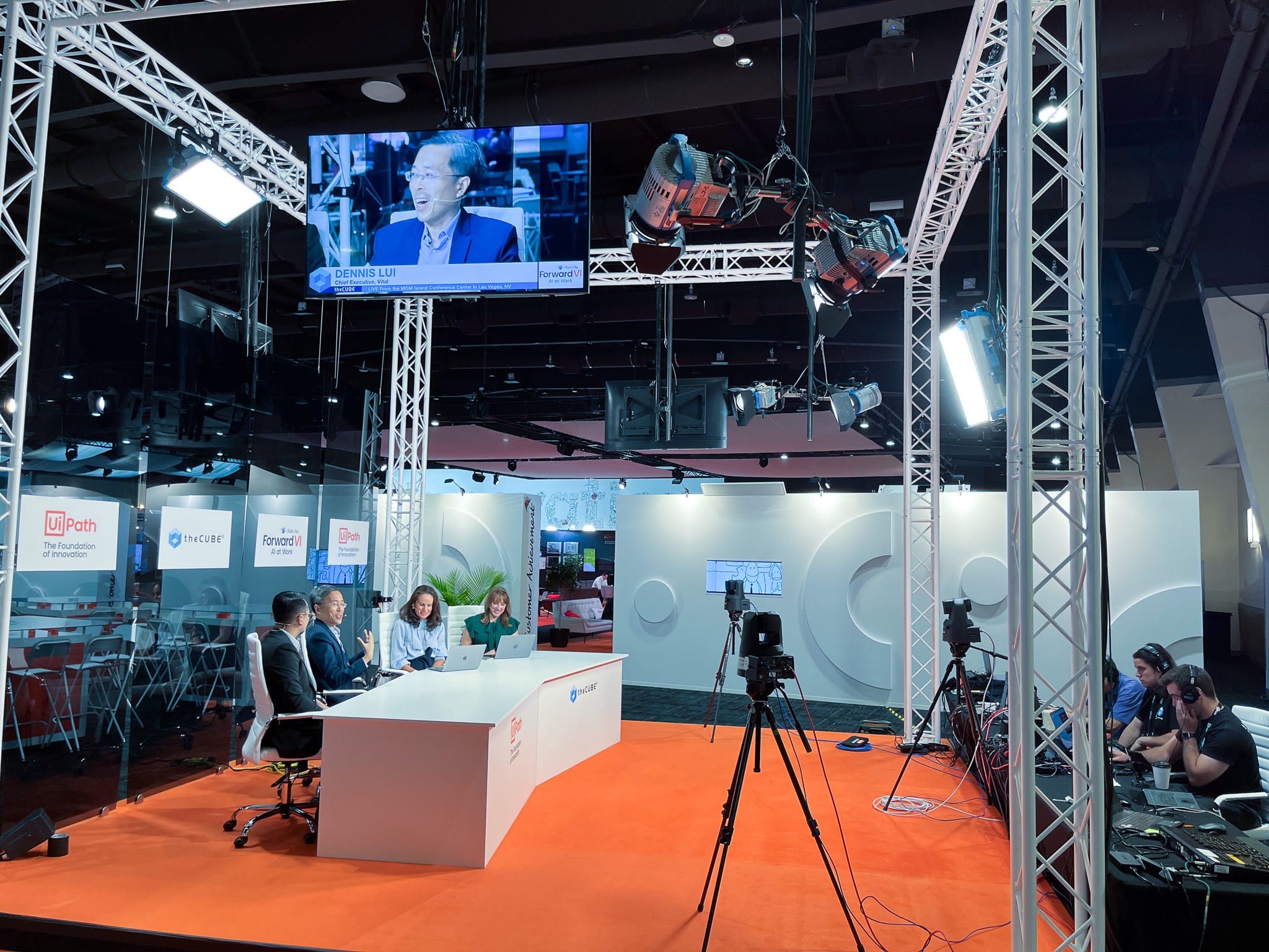 A live studio set with four people seated at a desk, cameras, studio lights, and a screen showing Dennis Lui being interviewed.
