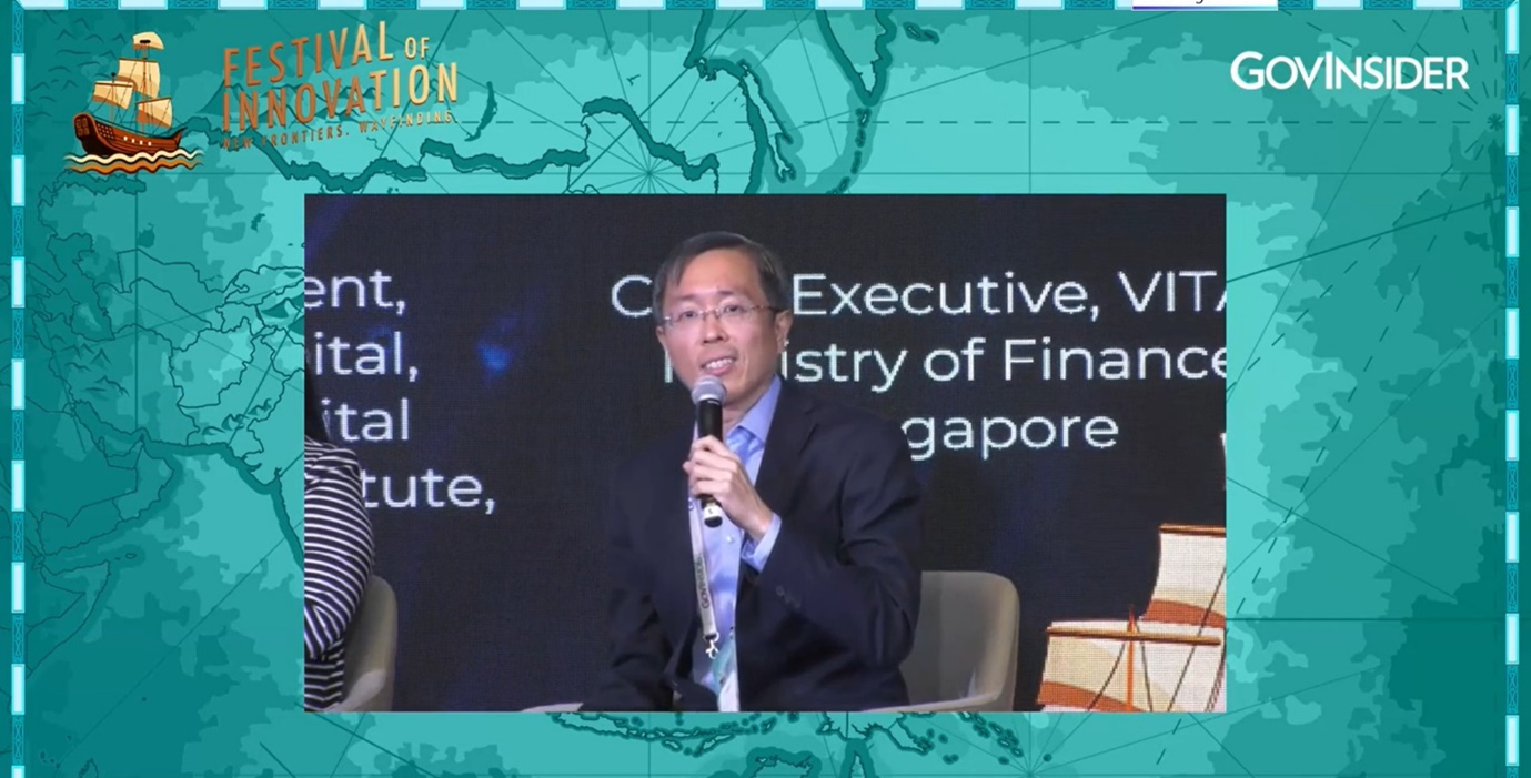 Man in a suit speaking into a microphone at GovInsider's Festival of Innovation, with a Ministry of Finance Singapore backdrop.