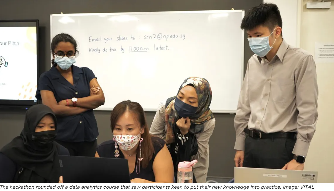 Five masked people gather around a laptop in a classroom with a whiteboard showing an email instruction.