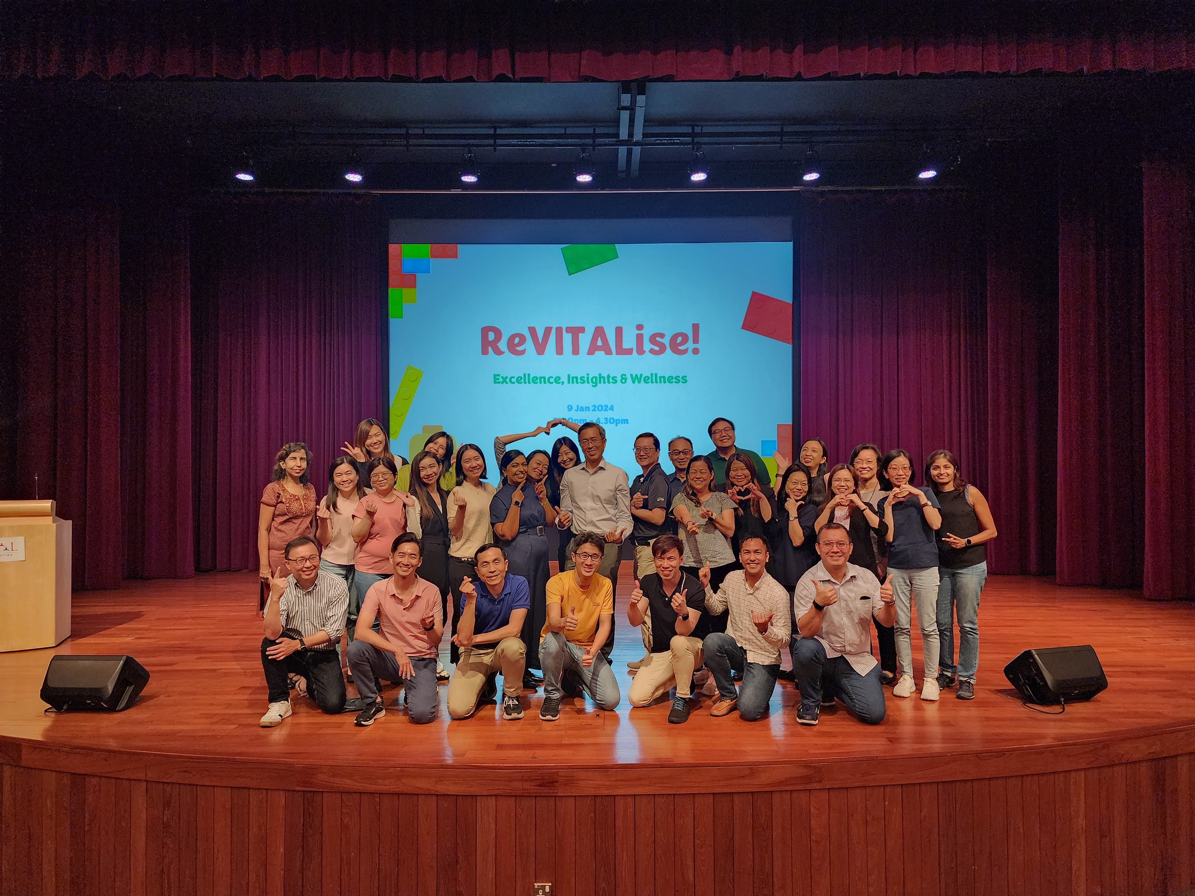 Group posing on auditorium stage before screen reading ReVITALise!; some kneel in front, purple curtains behind.