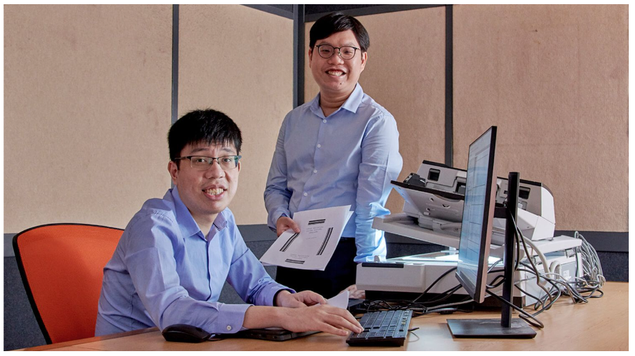 Two smiling men in blue shirts at a desk with a computer monitor, keyboard, and scanner in an office.