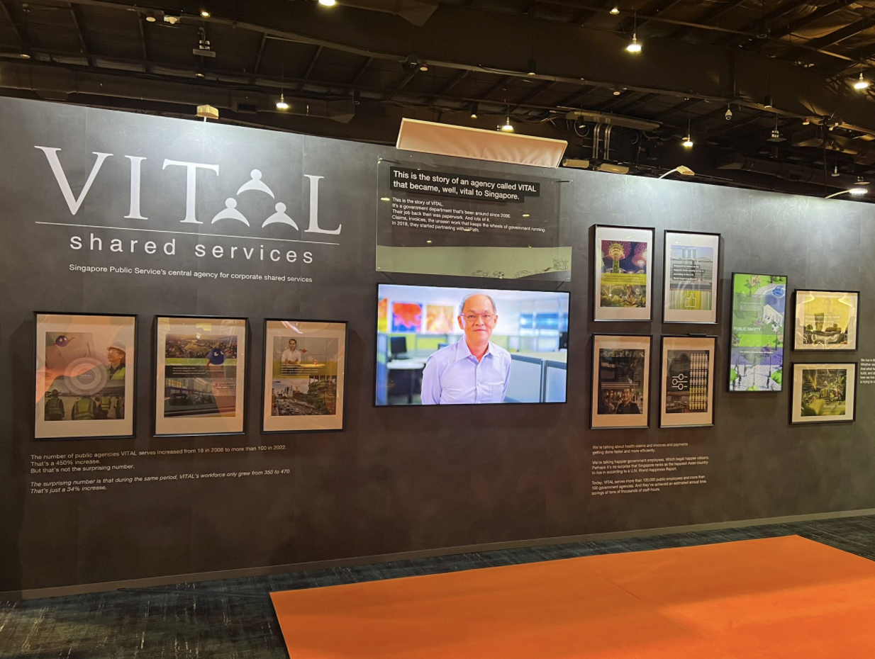 Dark exhibition wall displaying VITAL Shared Services branding, framed posters, a screen showing a man, and descriptive text panels.