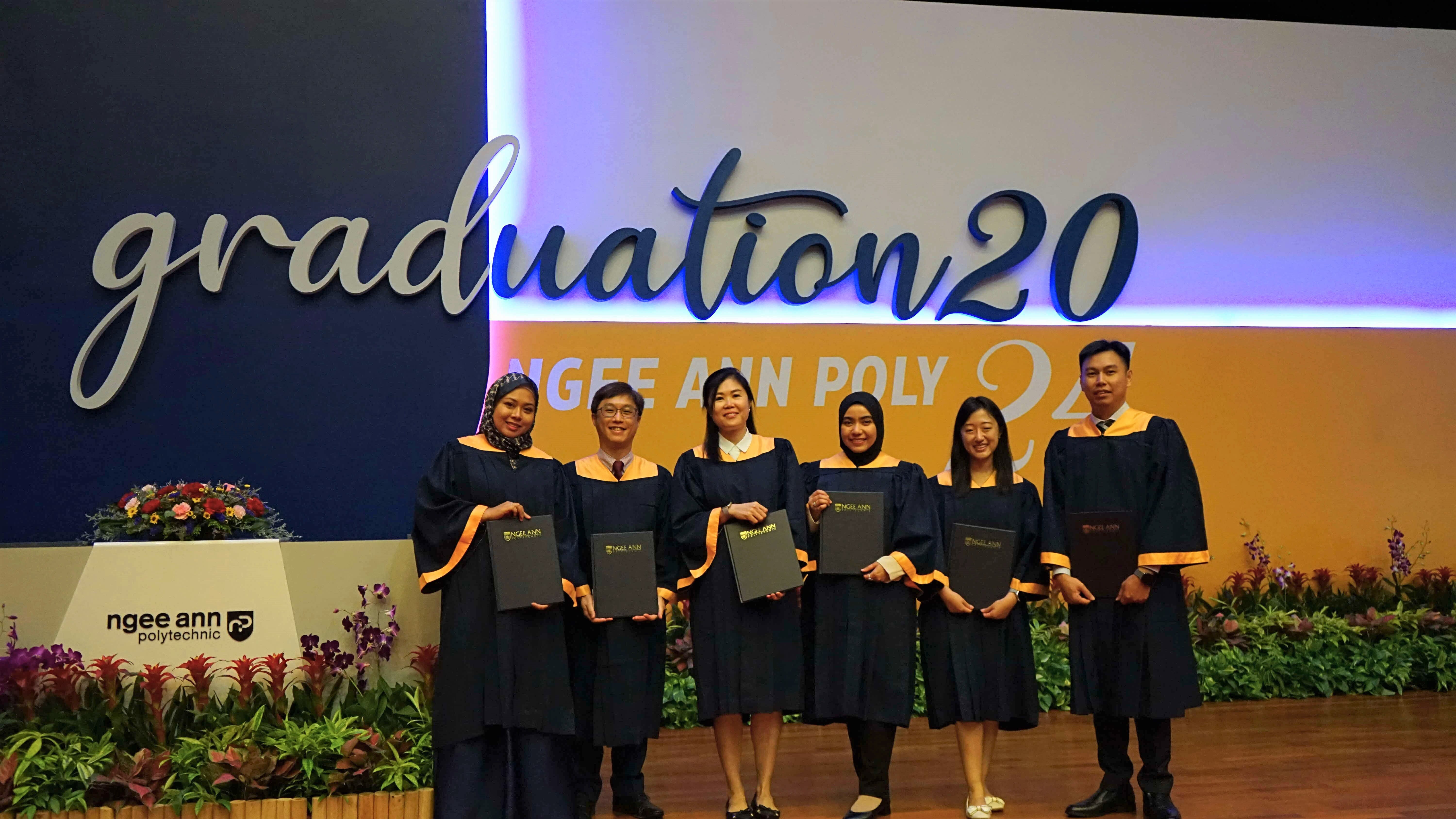 Six graduates in navy and gold robes hold diplomas on stage at Ngee Ann Polytechnic Graduation 2020.