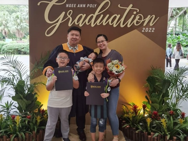 Family posing in front of Ngee Ann Poly Graduation 2022 backdrop, man in gown holding baby, two boys with folders.