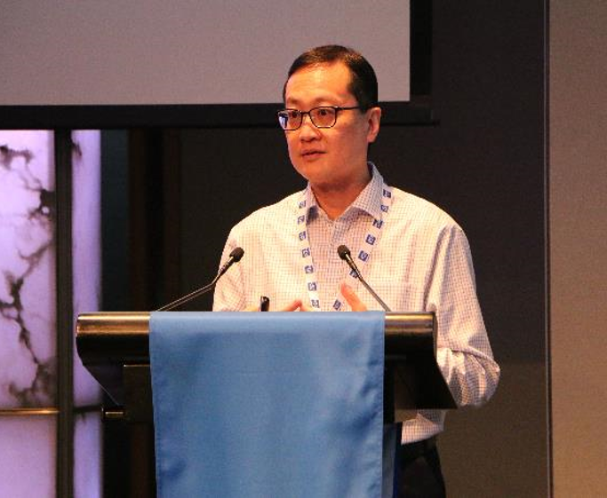 A man wearing glasses and a checkered shirt speaks at a podium with two microphones and a blue drape.