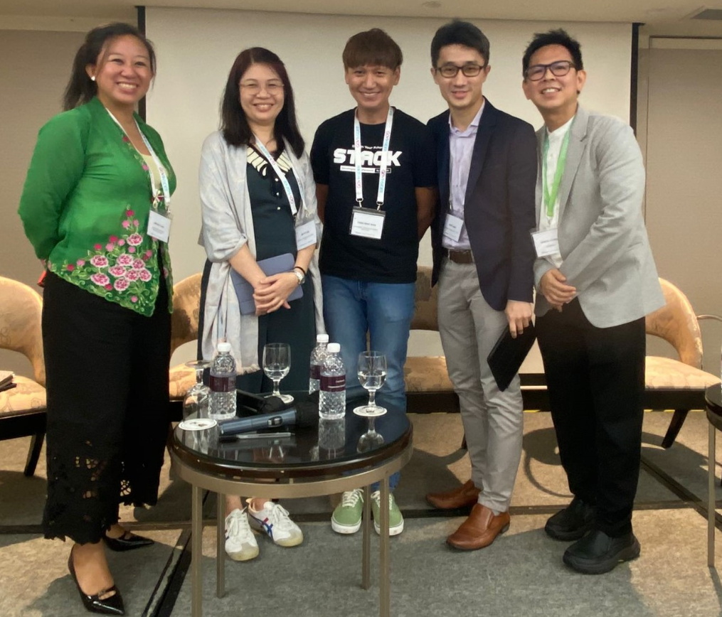 Five smiling conference attendees wearing lanyards pose together near a small round table with water bottles and glasses.